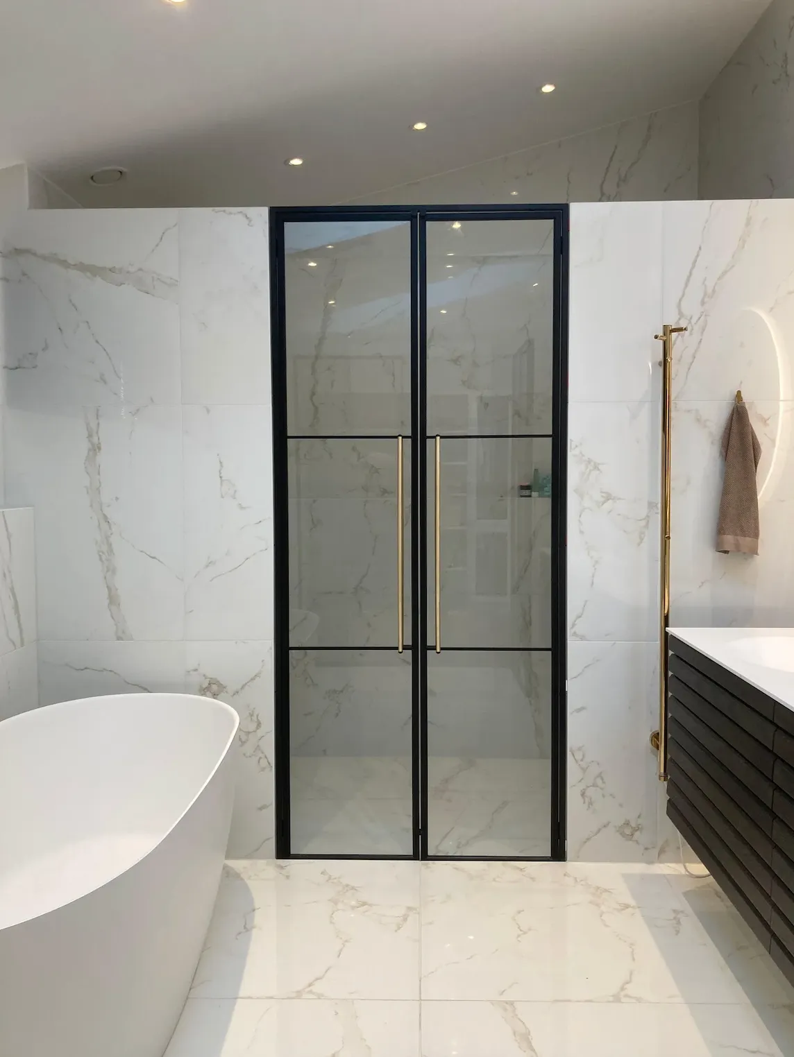 Moderne badeværelse med fritstående hvidt badekar, sort-ramme glasbrusedør og marmorerede vægge og gulv.