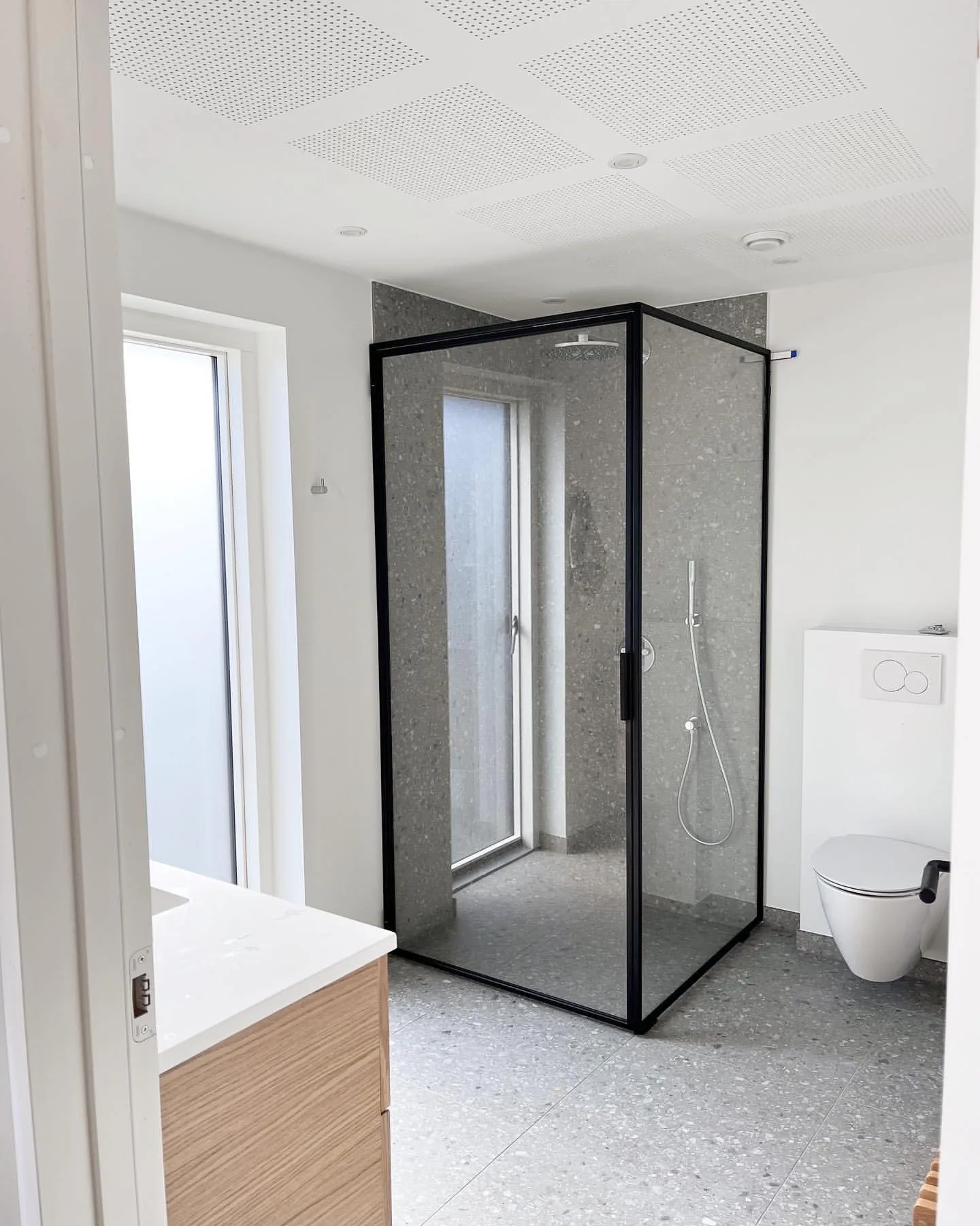 Moderne badeværelse med gennemsigtig bruseniche med sort ramme, væghængt toilet og træskab med hvid bordplade.