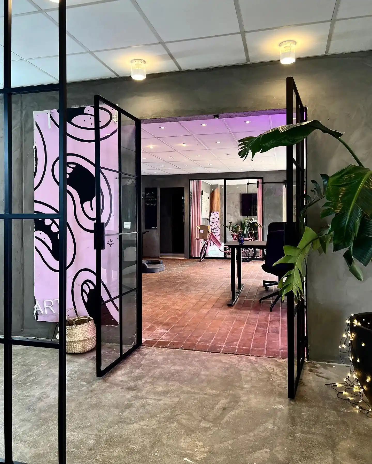 Indgang til moderne kontor med sort metalrammedøre, stor grøn plante og cykel i rummeligt rum med lyserød lys.