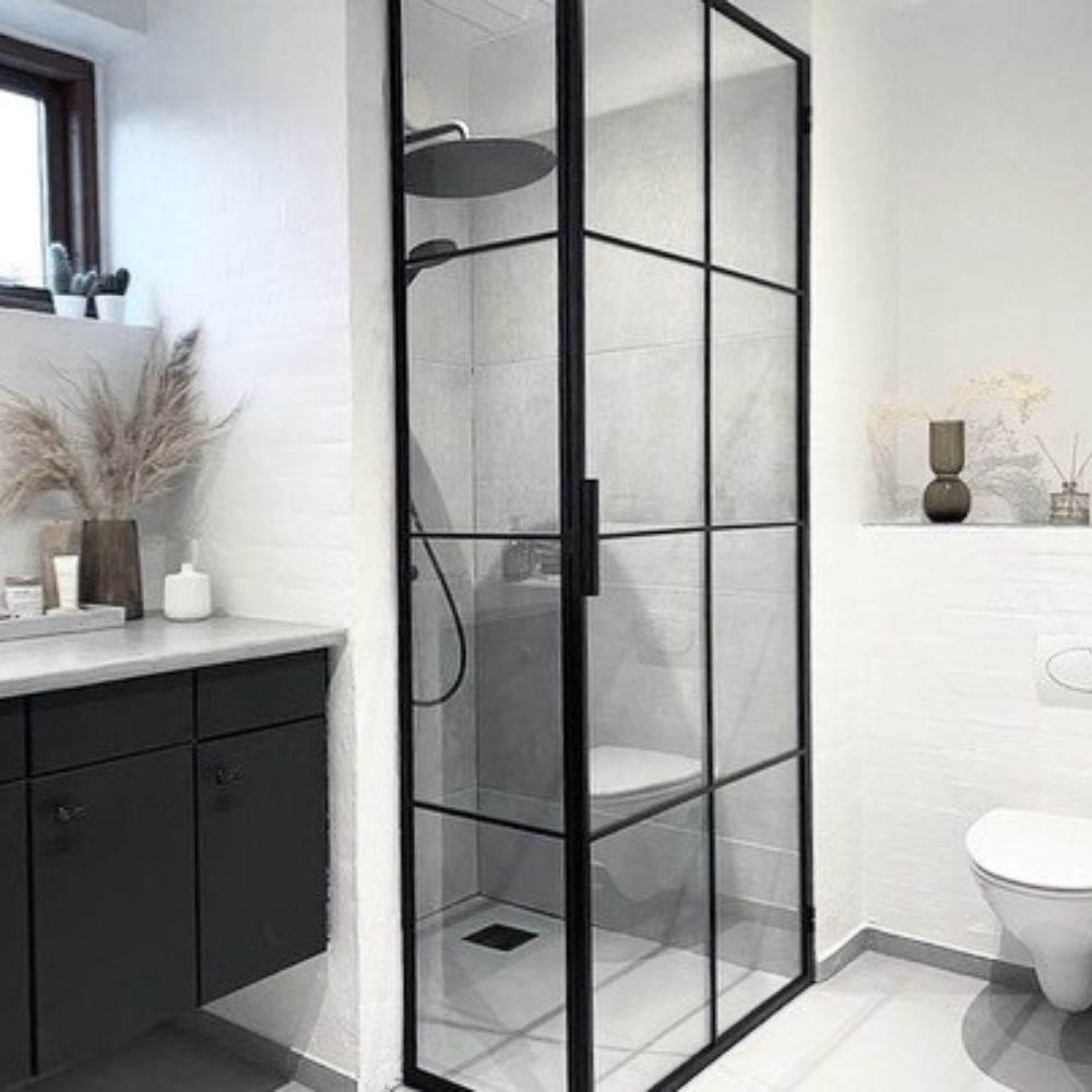 Moderne badeværelse med sortrammet glasbrusekabine, hvidt toilet, sort skab og dekorative planter.
