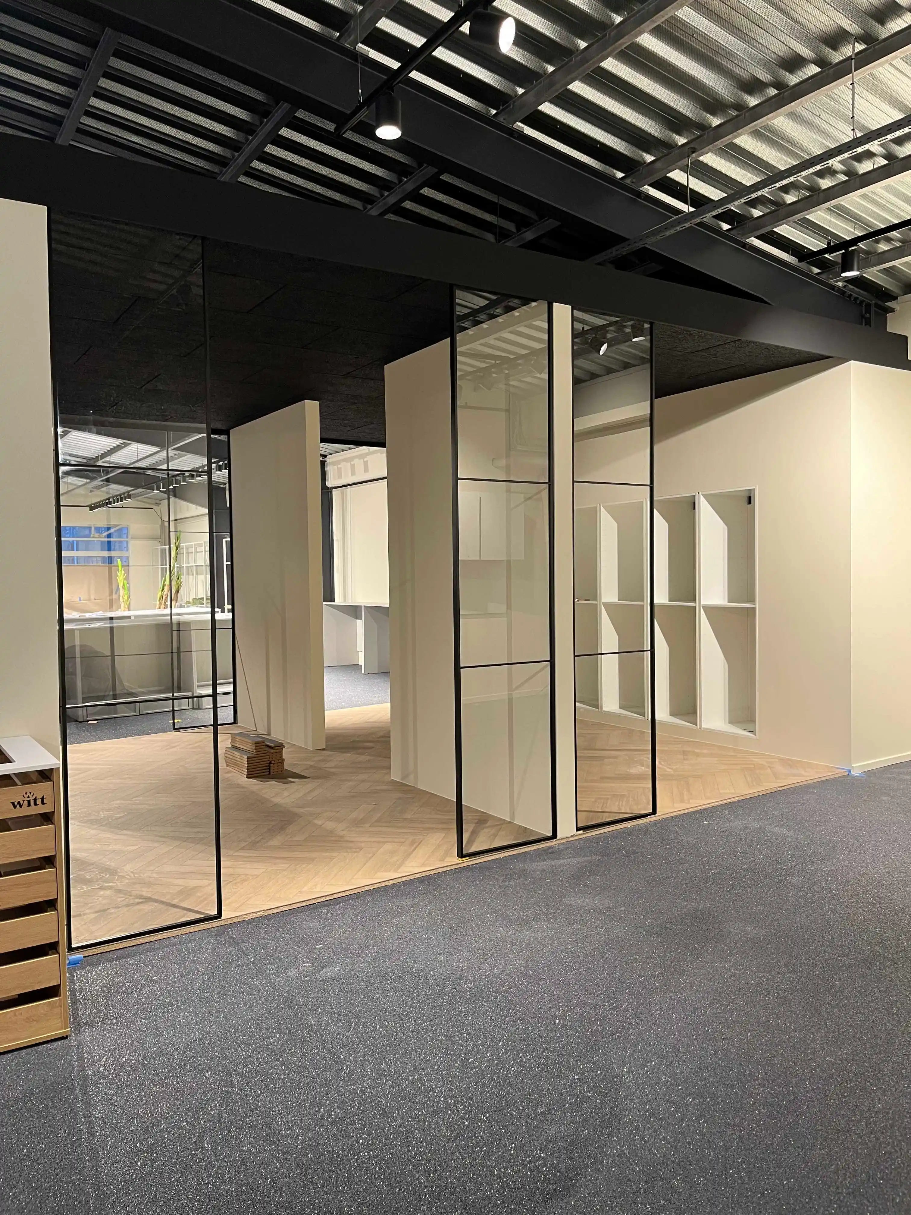 Moderne kontorlokale med glasvægge, trægulv og åben loftkonstruktion med indbyggede spotlys.