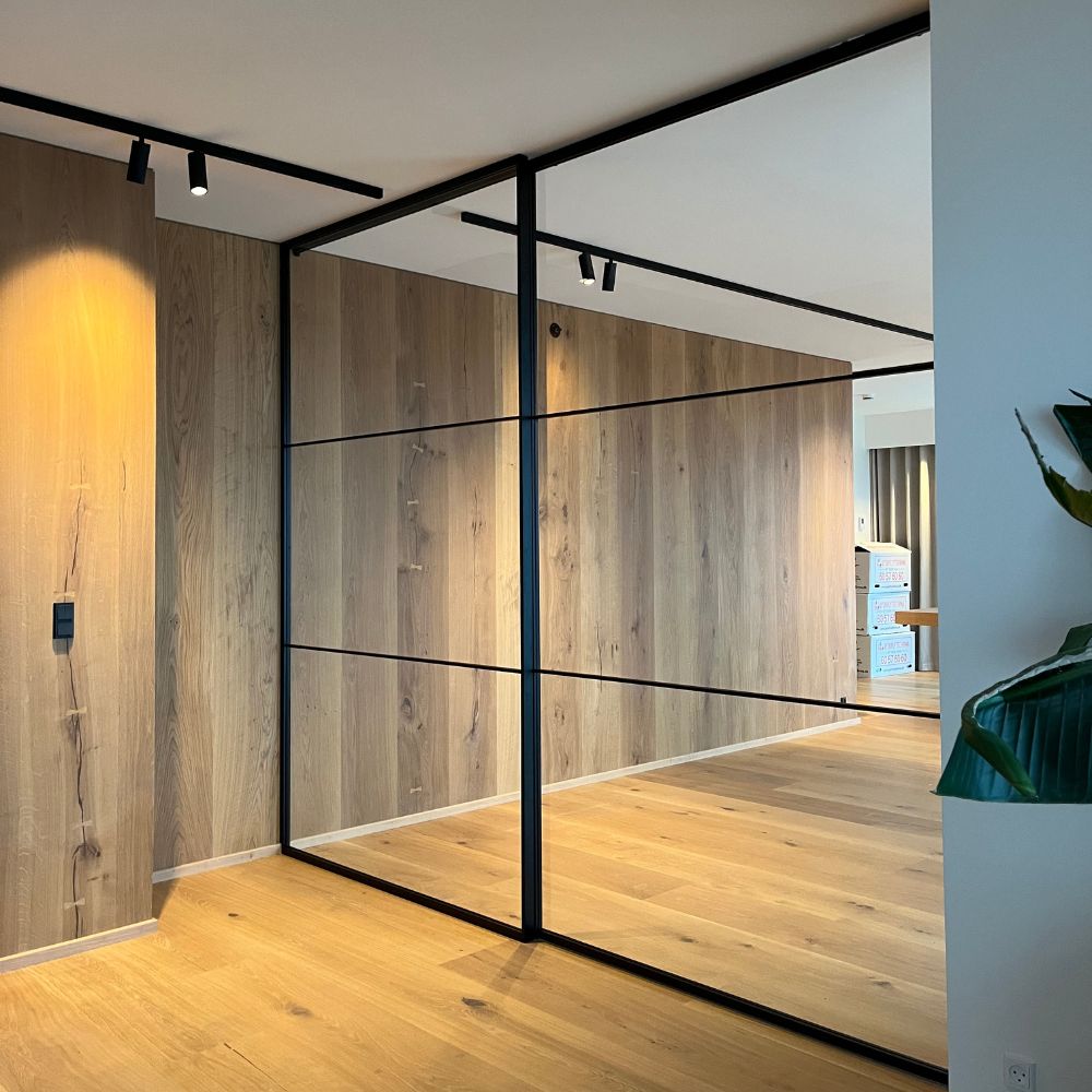 Moderne rum med lyst trægulv, vægbeklædning i træ, og en stor glasvæg med sort ramme.