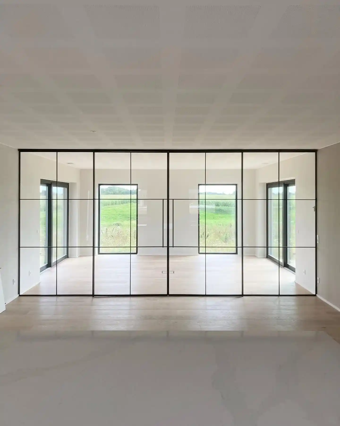 Moderne tomt rum med store glasvægsdøre med sorte rammer, der åbner mod en grøn natur.