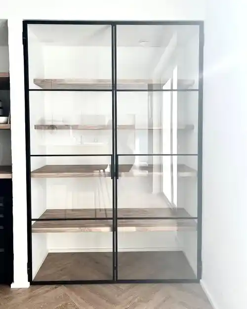 Glasvitrineskab med sorte rammer og hylder i lyst træ i et lyst rum.