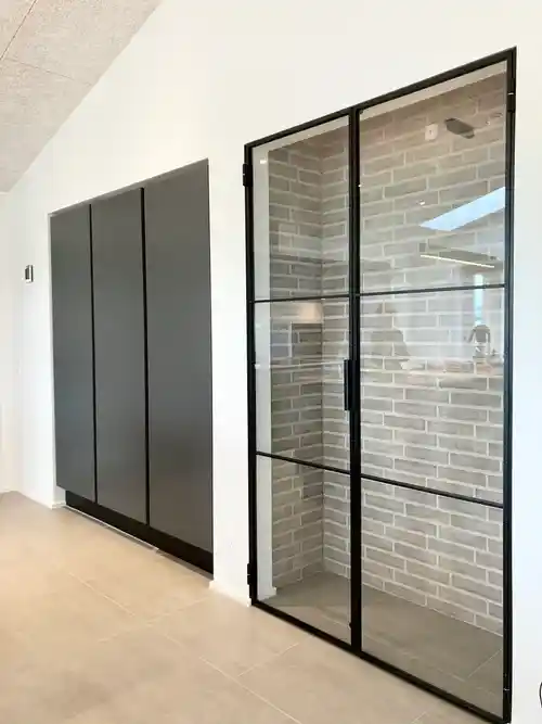 Moderne gang med mørke skabe og glasdør med sort ramme, der viser væg med grå mursten bagved.