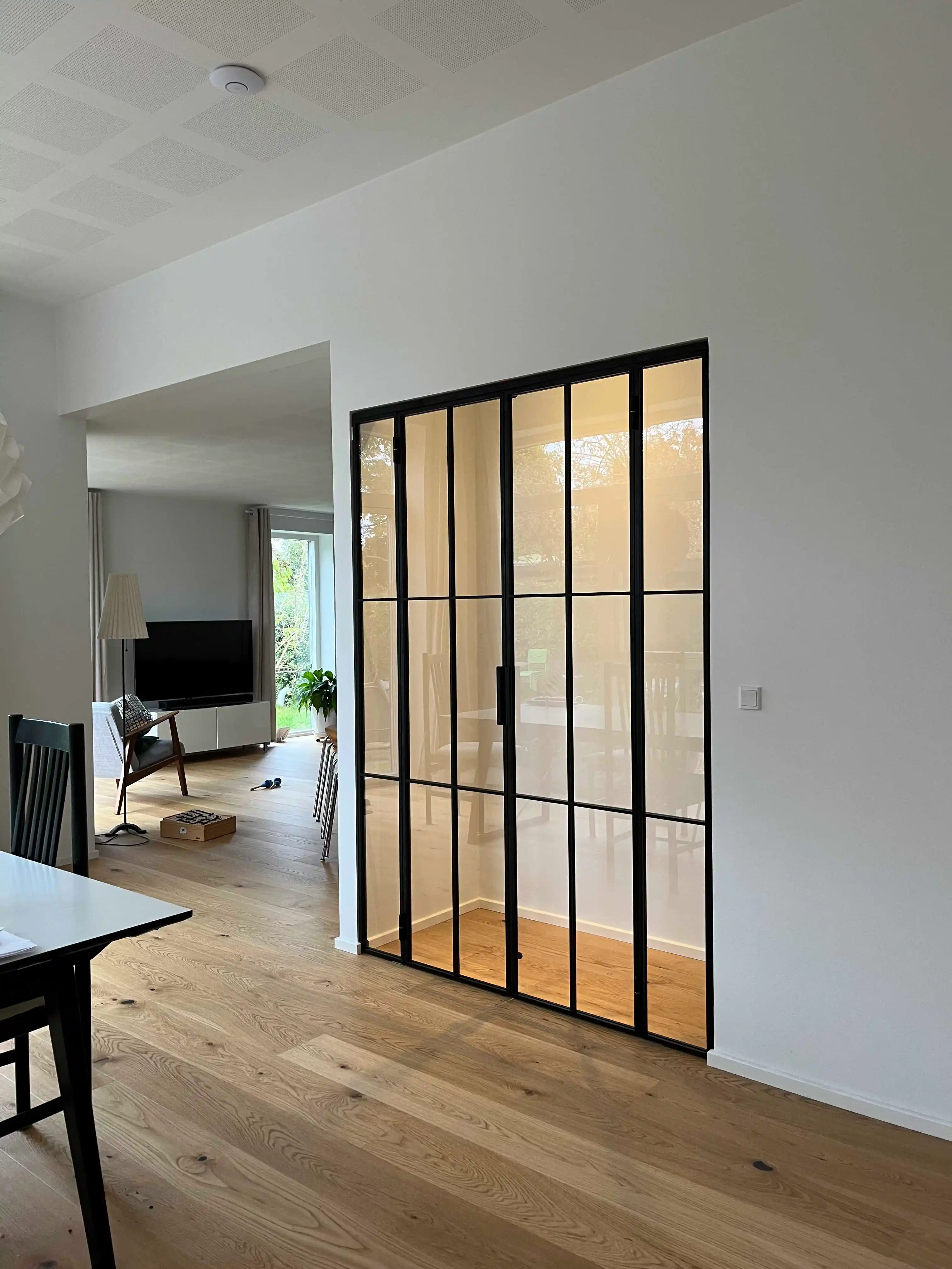 Moderne stue med glasdør med sort ramme, lyst trægulv og fjernsyn på skænk i baggrunden.