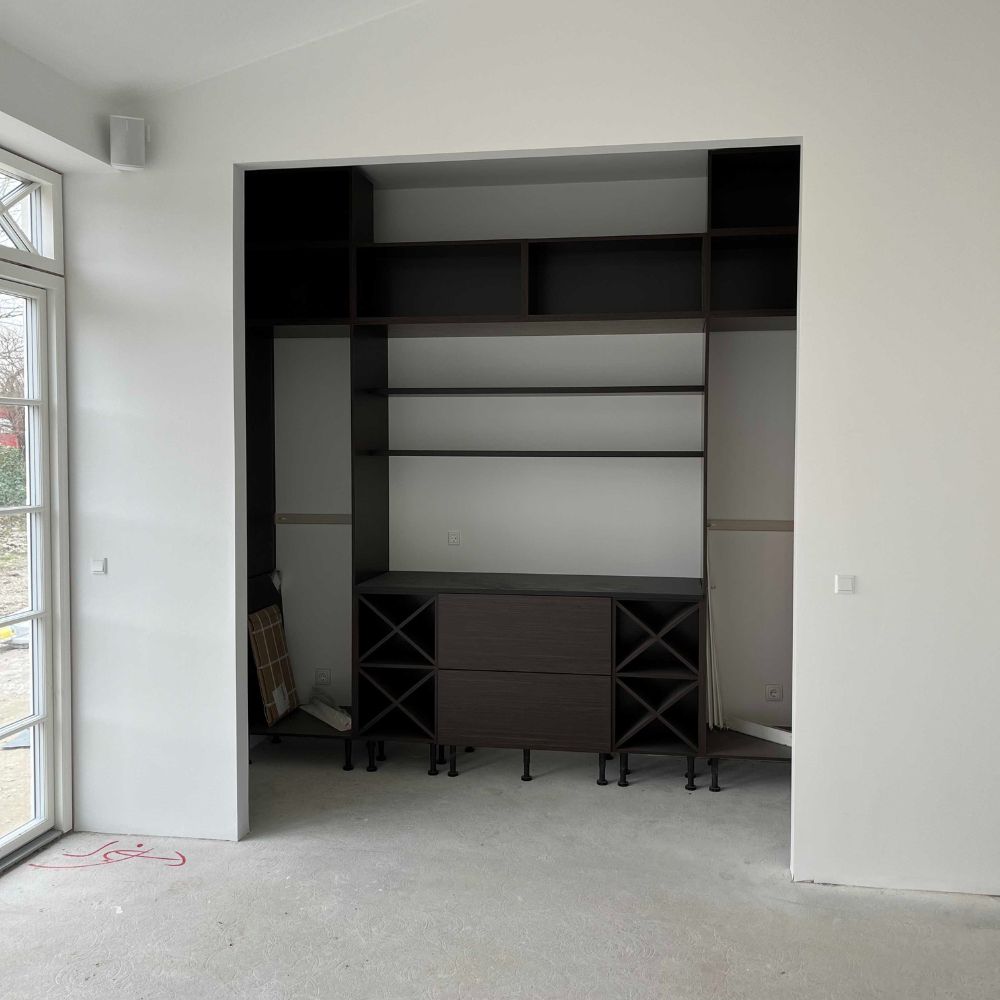 Sort, tomt vægindbygget reolsystem med hylder og skuffer i et rum med hvide vægge og et stort vindue til venstre.