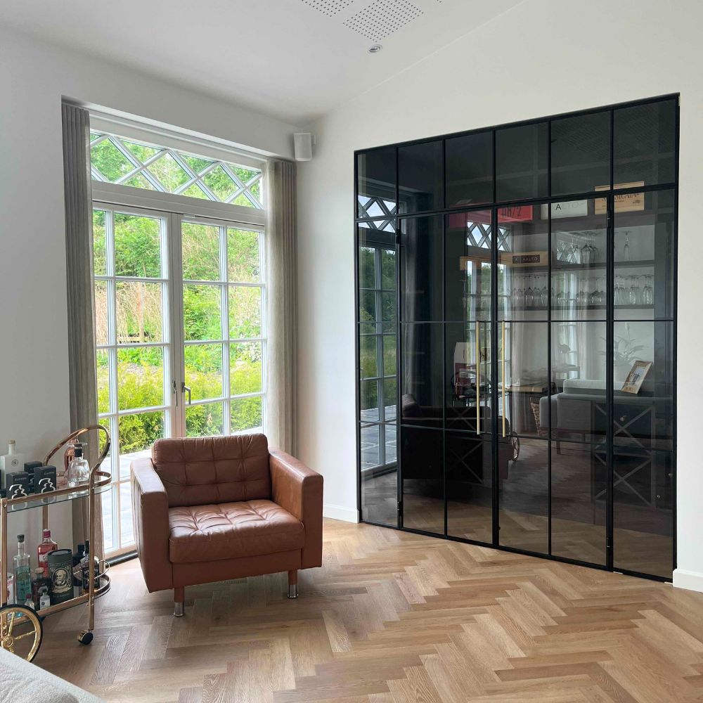 Lyst rum med brunt læderlænestol ved store hvidrammede vinduer og sorte glasdøre med metalramme.