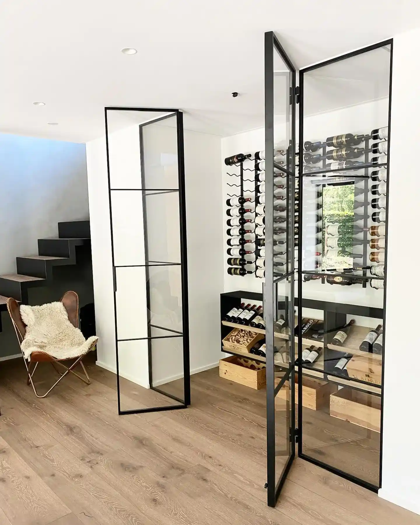Moderne vinkælder med glasdøre, vægmonterede vinholdere, trævinbokse og en lænestol med plyssoverflade foran trappe.