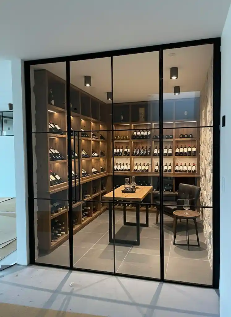 Moderne vinkælder med træreoler fyldt med vinflasker, et bord med karafler og et læderstol med et lille rundt bord.