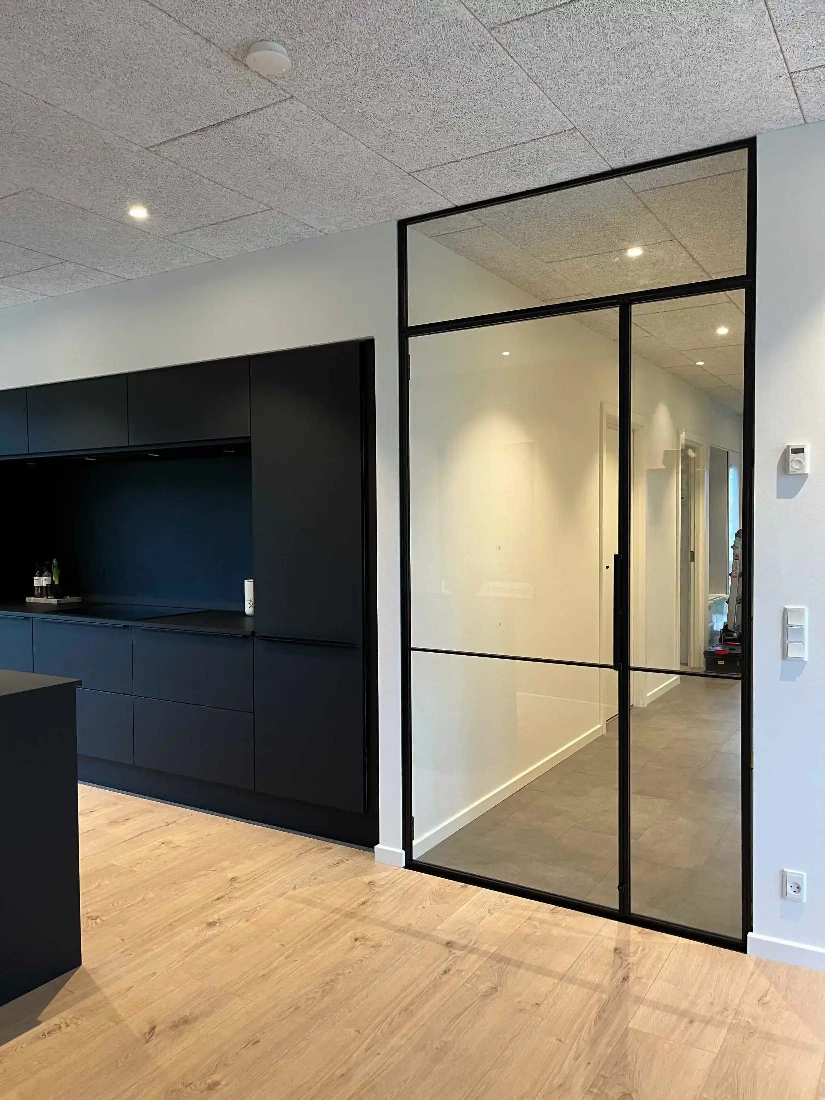 Moderne køkken med sorte skabe og glasdør med sort ramme, der fører til en gang med gråt gulv.