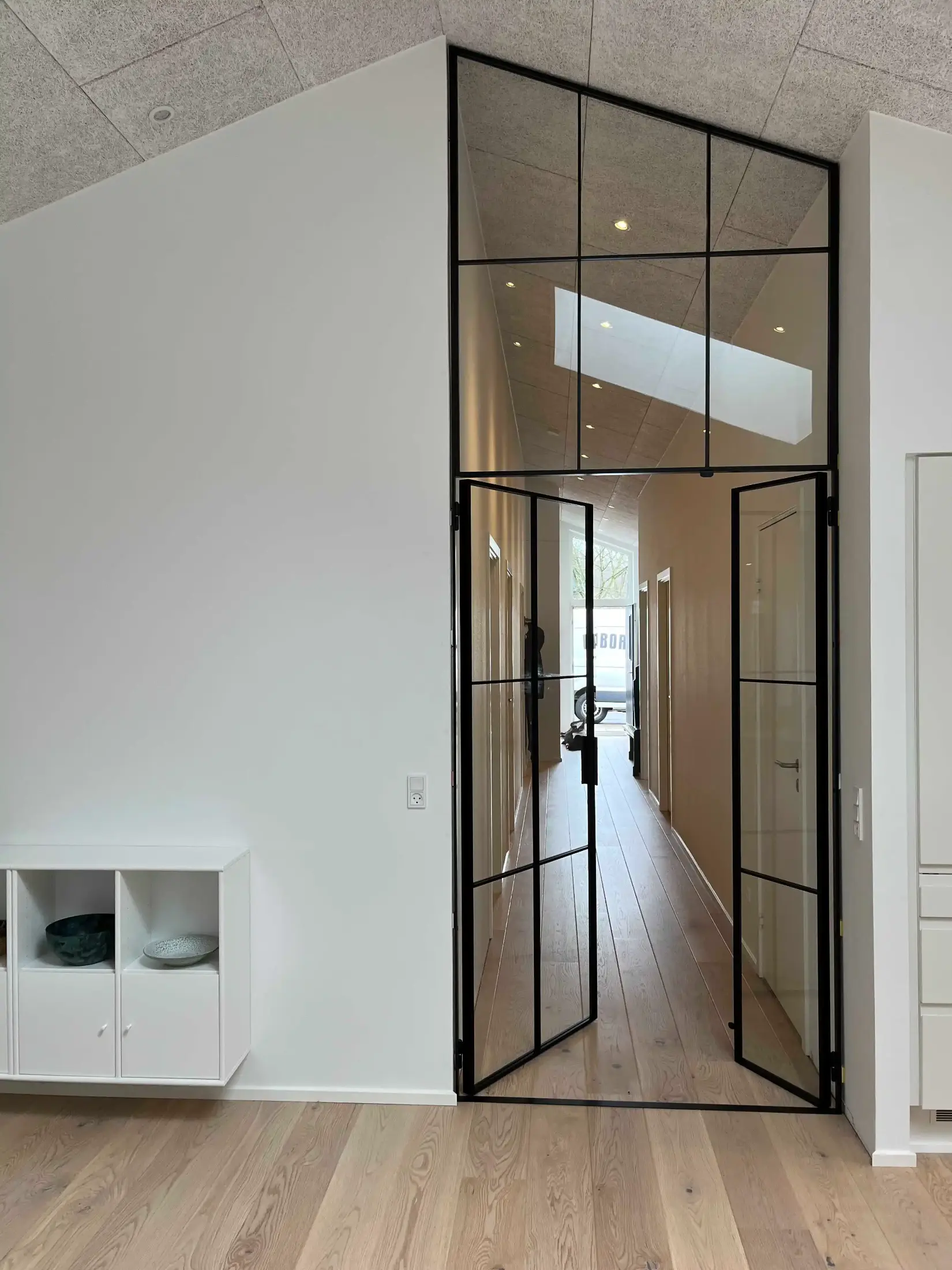 Lang gang med hvidmalede vægge, lyst trægulv og sorte, rammelagte glasdøre med ovenlysvindue i et moderne hjem.