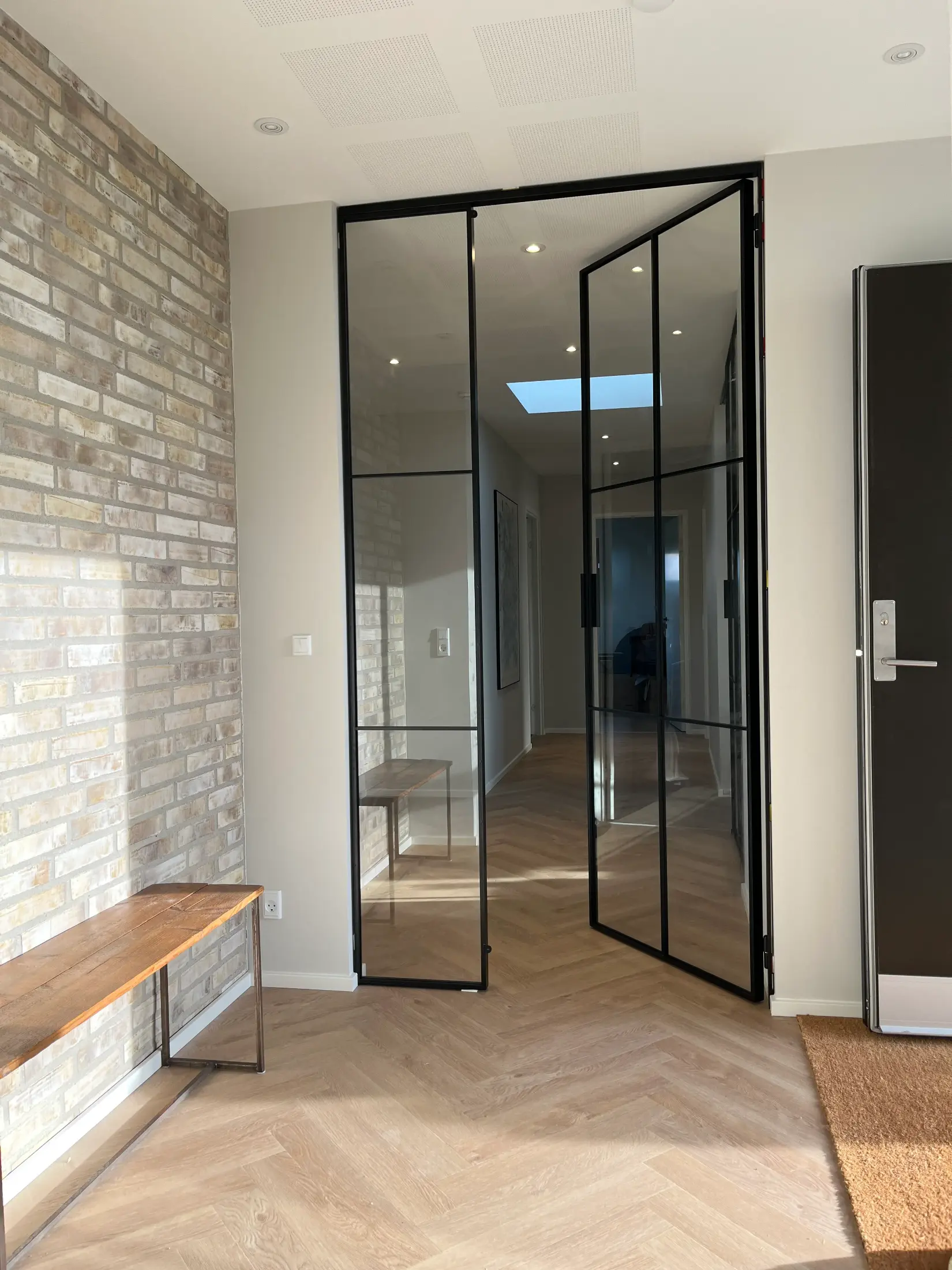 Indgang med lyst trægulv i sildebensmønster, murstensvæg, træbænk og sorte glasdøre, der åbner til en lys gang med loftslys.