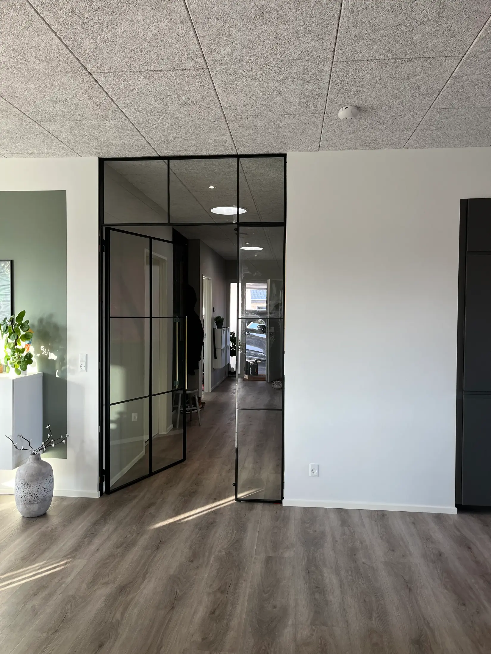 Moderne entré med glasdør i sort metalramme, lyst trægulv og hvide vægge.