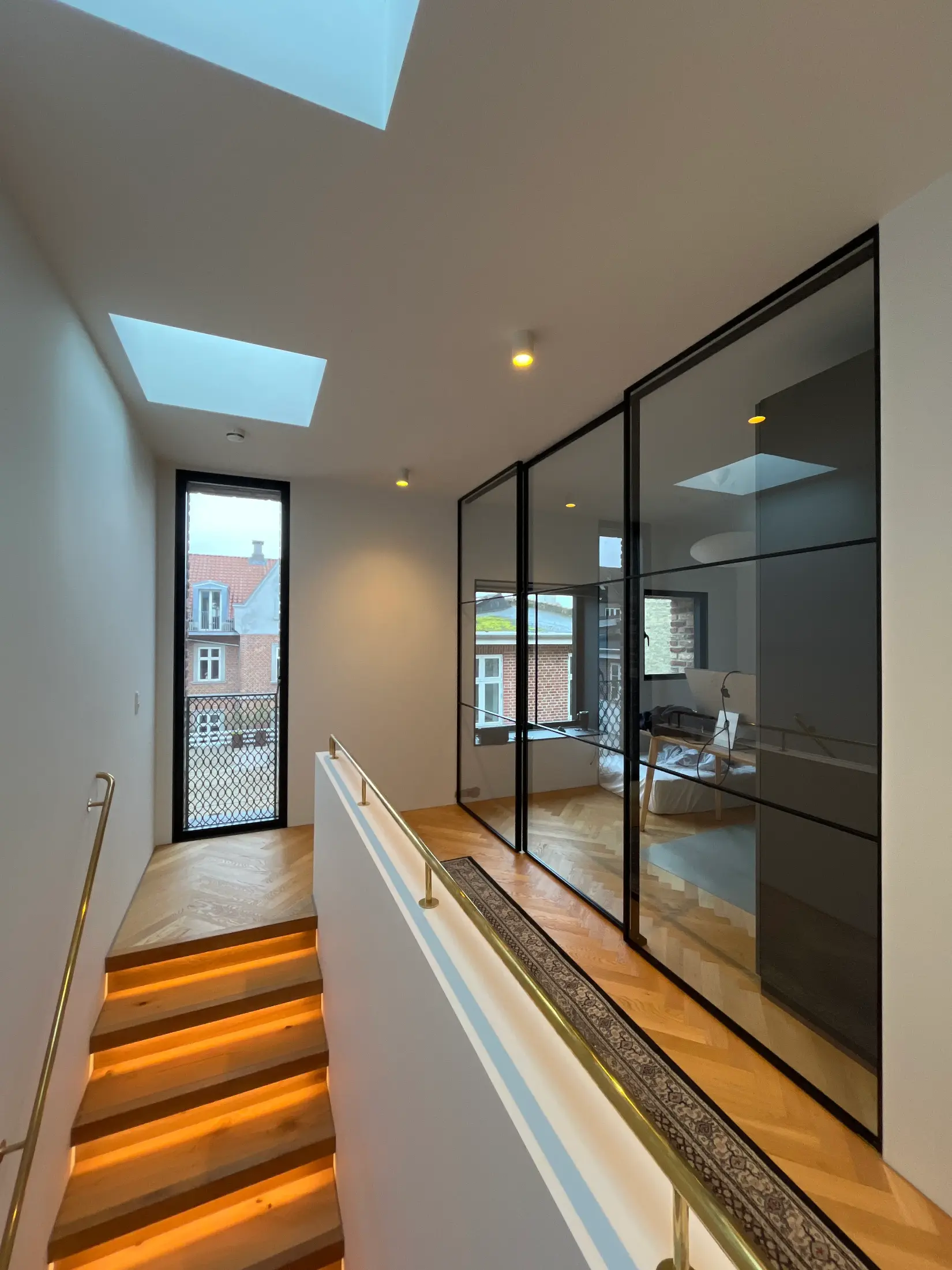 Moderne trappe med indbygget LED-belysning, glasvægge og højt vindue med udsigt til byen.