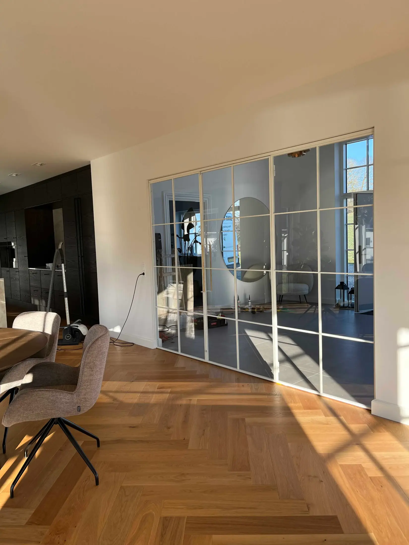 Moderne stue med trægulv, runde spejl på væggen bag glasdøre og grå stole ved et rundt bord.