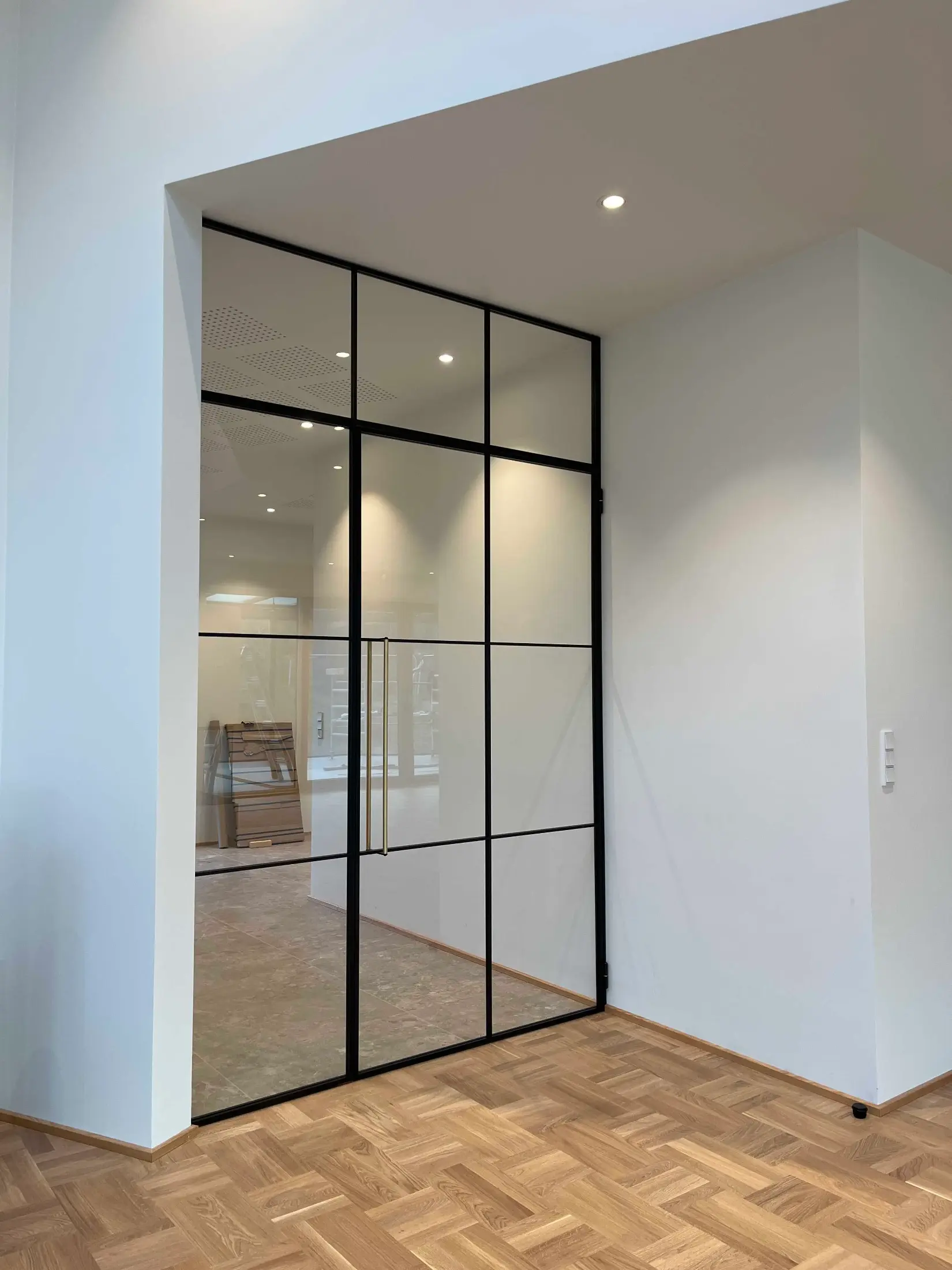 Moderne skydedør med sort ramme og glas mellem to hvide vægge og lyst parketgulv.
