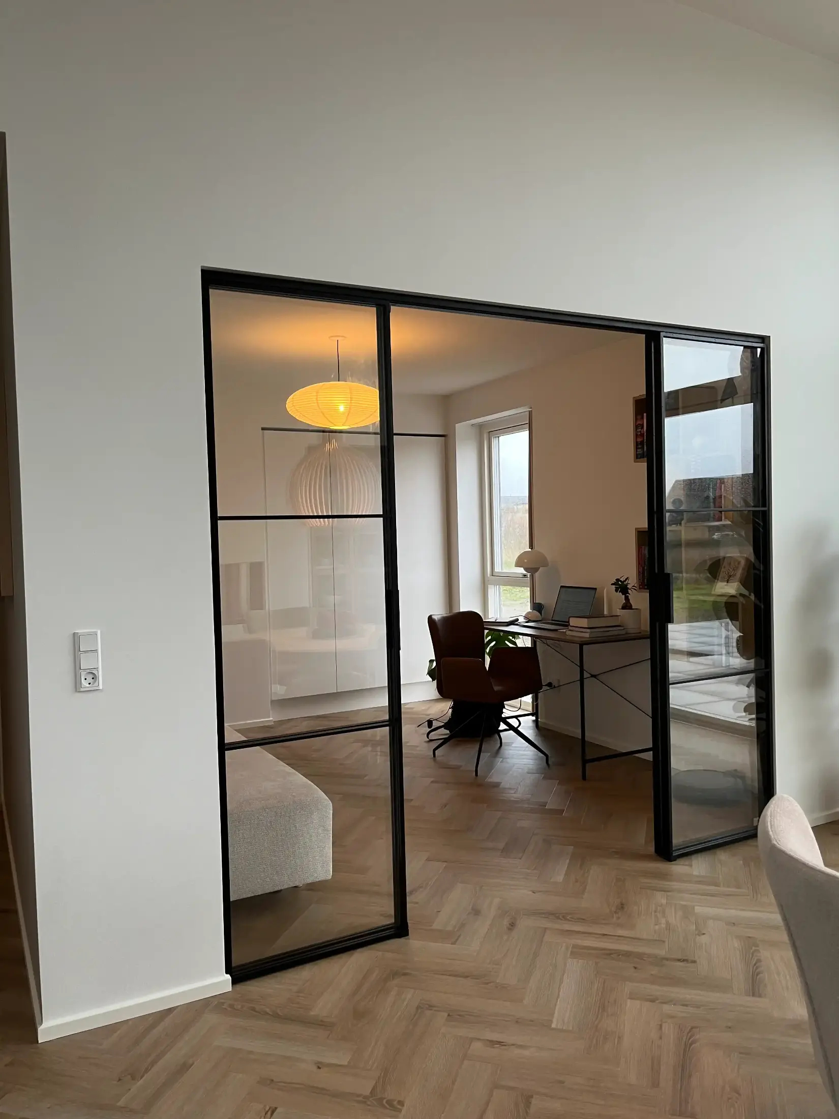 Indendørs udsigt til et moderne hjemmekontor med et brunt kontorstol, skrivebord med åben bærbar computer og store vinduer, adskilt af glasdøre med sorte rammer.