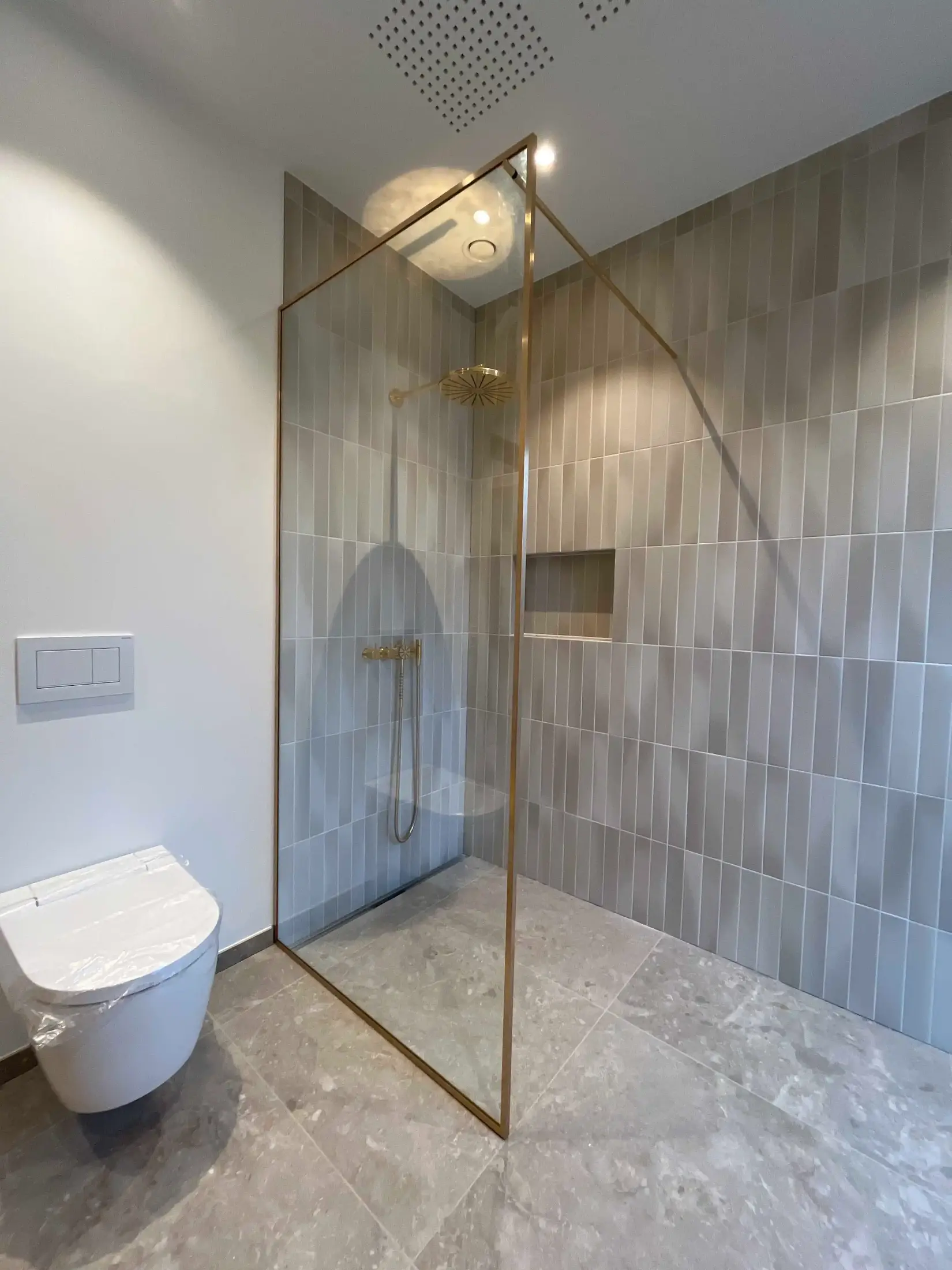 Moderne badeværelse med glasbruser med guldkant, grå fliser på væggen og væghængt toilet.