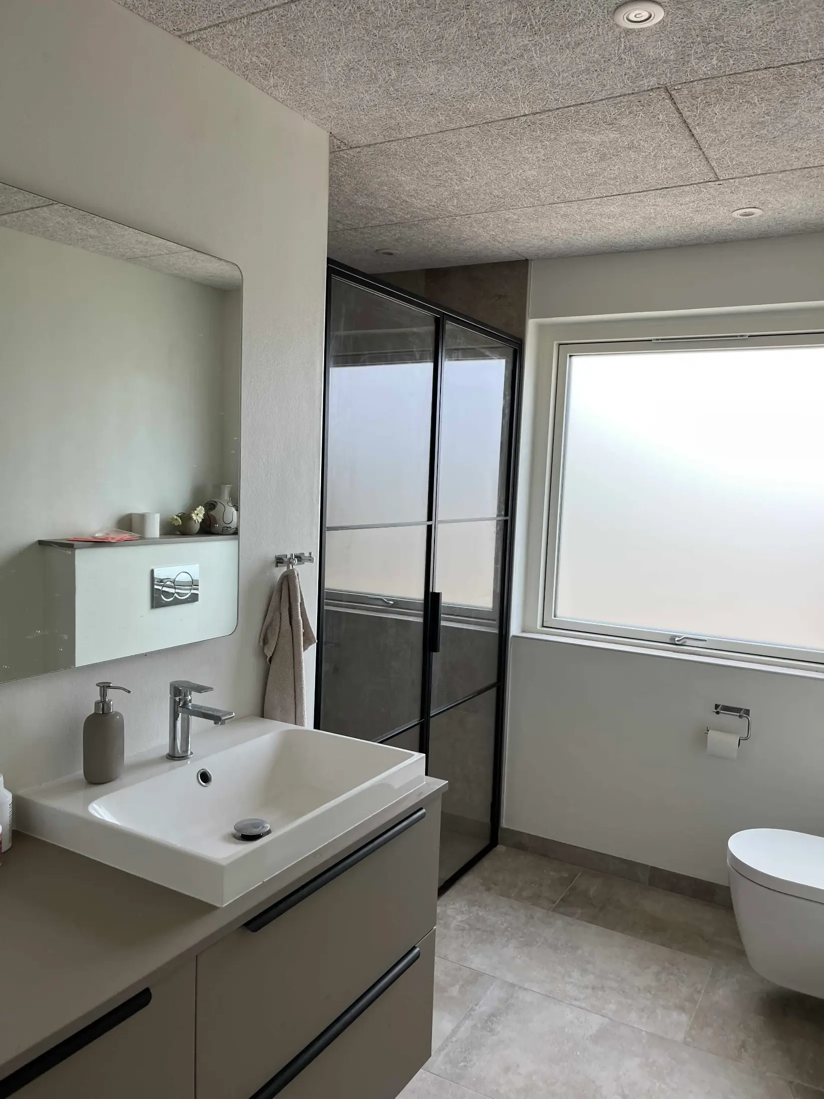 Moderne badeværelse med vask med hvid skål, spejl, håndklæde, glasbruser og væghængt toilet ved stort frostet vindue.