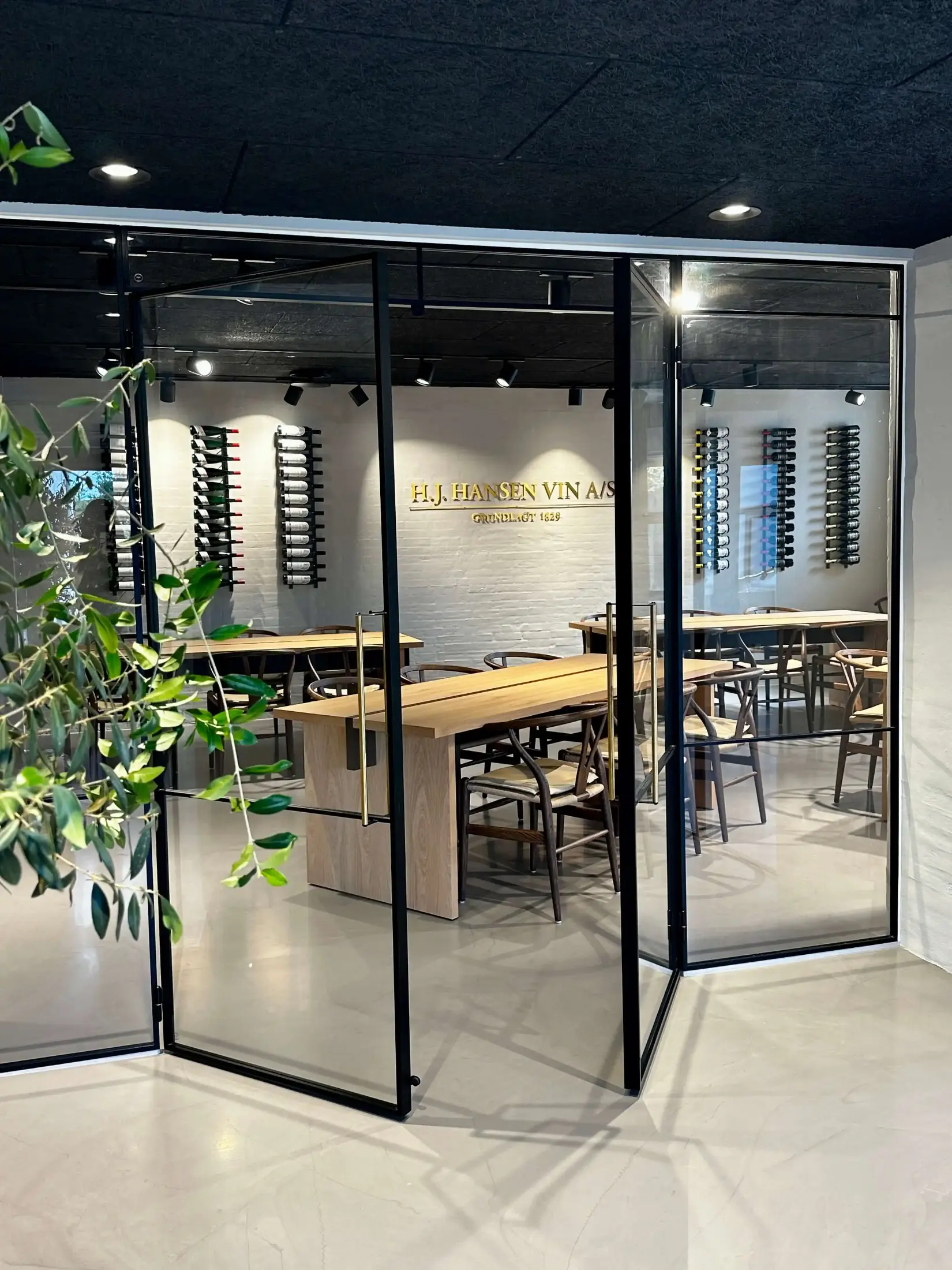 Mødelokale med glasvægge, træborde og stole, vinflasker på vægmonterede stativer og H.J. Hansen Vin A/S logo på hvid murstensvæg.
