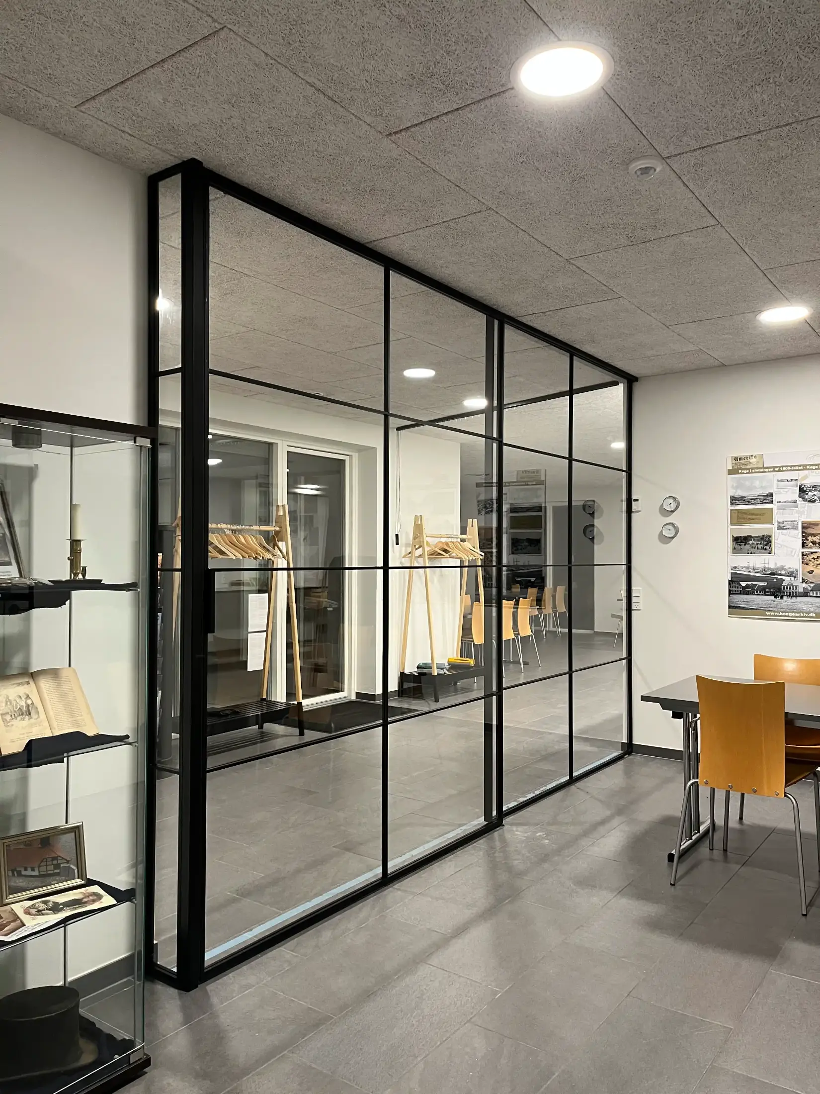 Moderne kontorindretning med glasvæg i sort ramme, træstole omkring et bord og en reol med bøger og billeder.