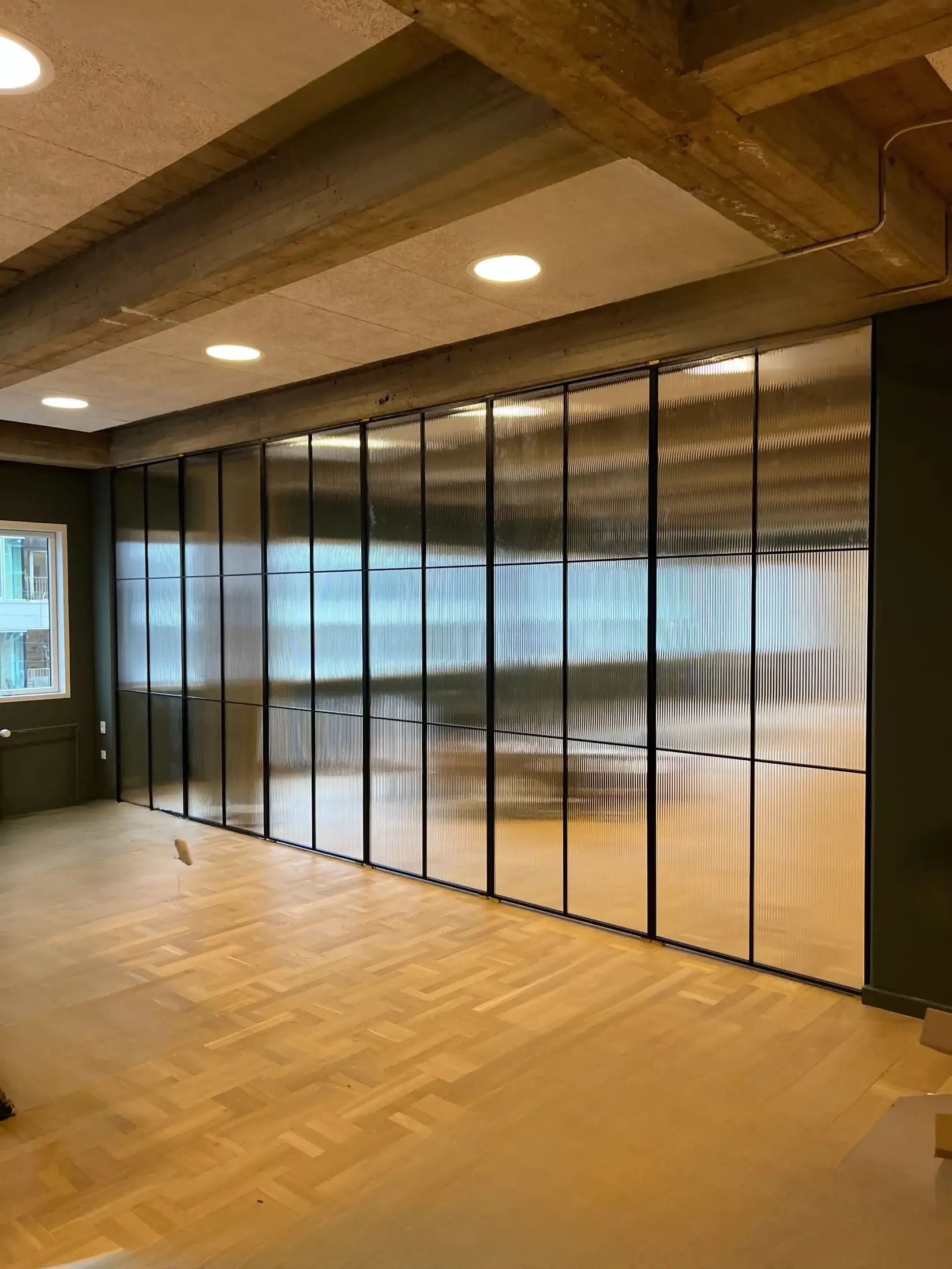 Moderne tomt rum med trægulv, store rippede glasvægge i sort ramme og indbyggede loftslamper.