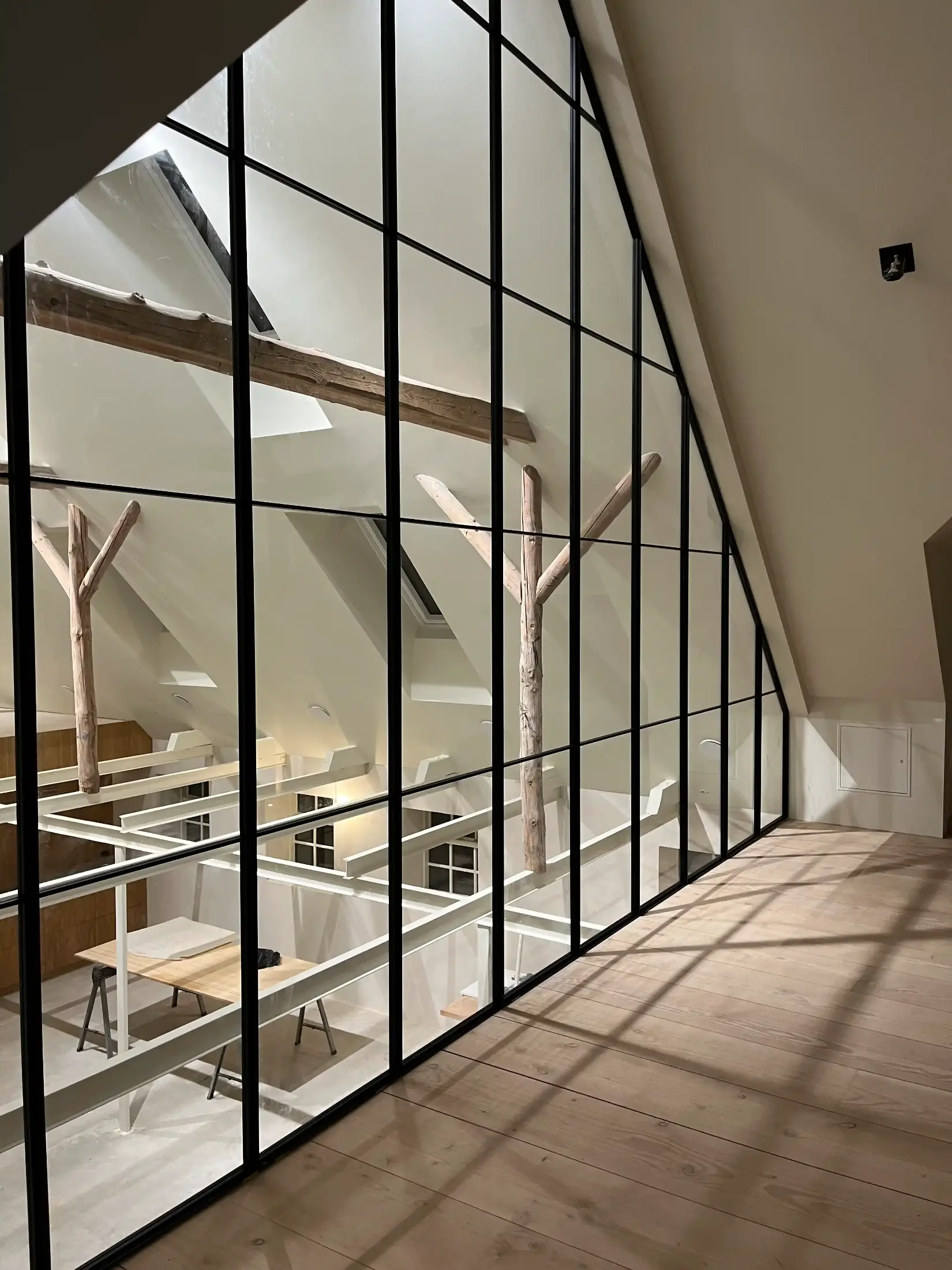 Loftkontor med store glasvægge, træbjælker og lyse trægulve, der adskiller arbejdsområder.