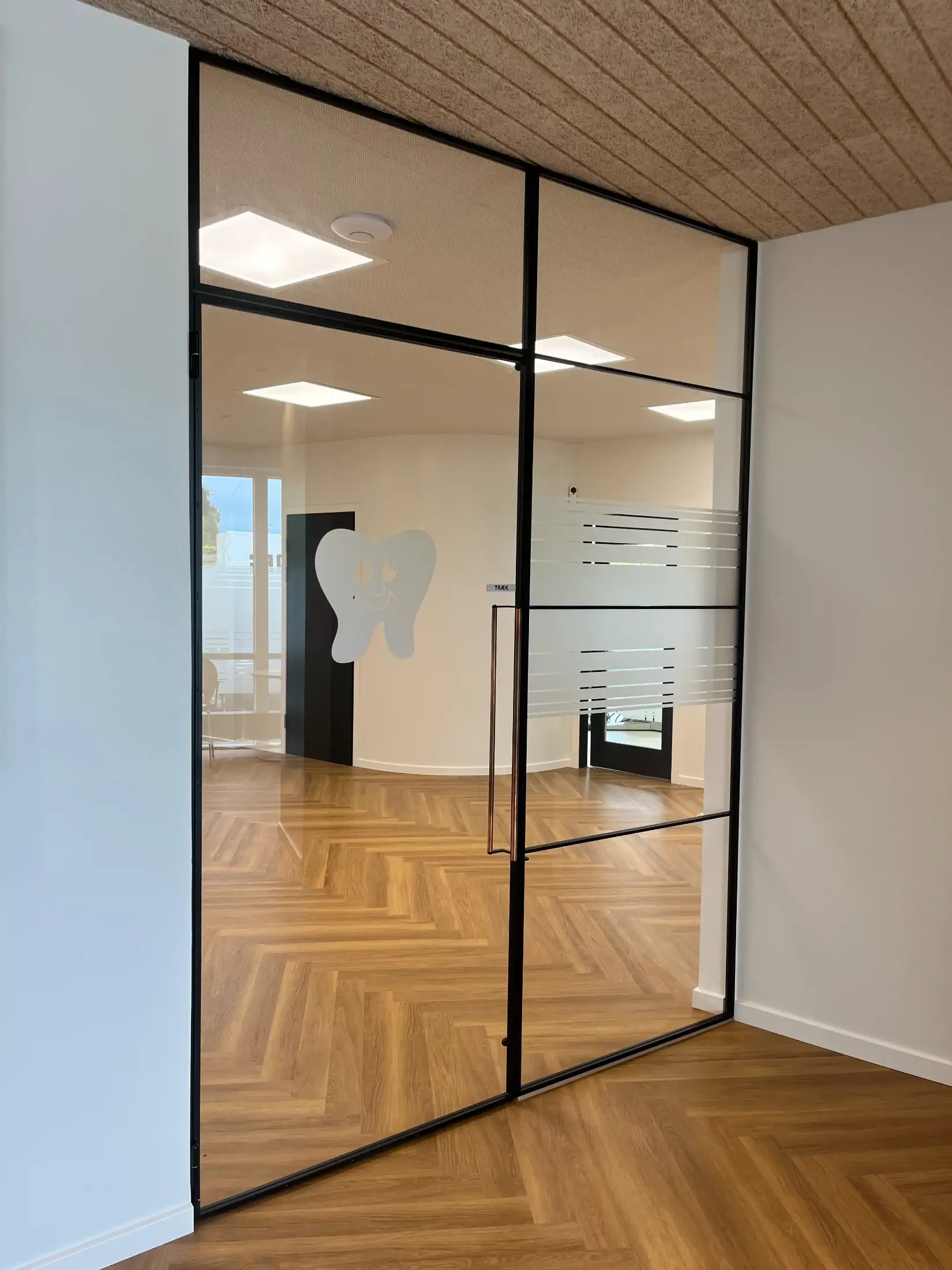 Glasdør med sort metalramme og et hvidt tandlogo, der fører ind til et lyst rum med trægulv i sildebensmønster.
