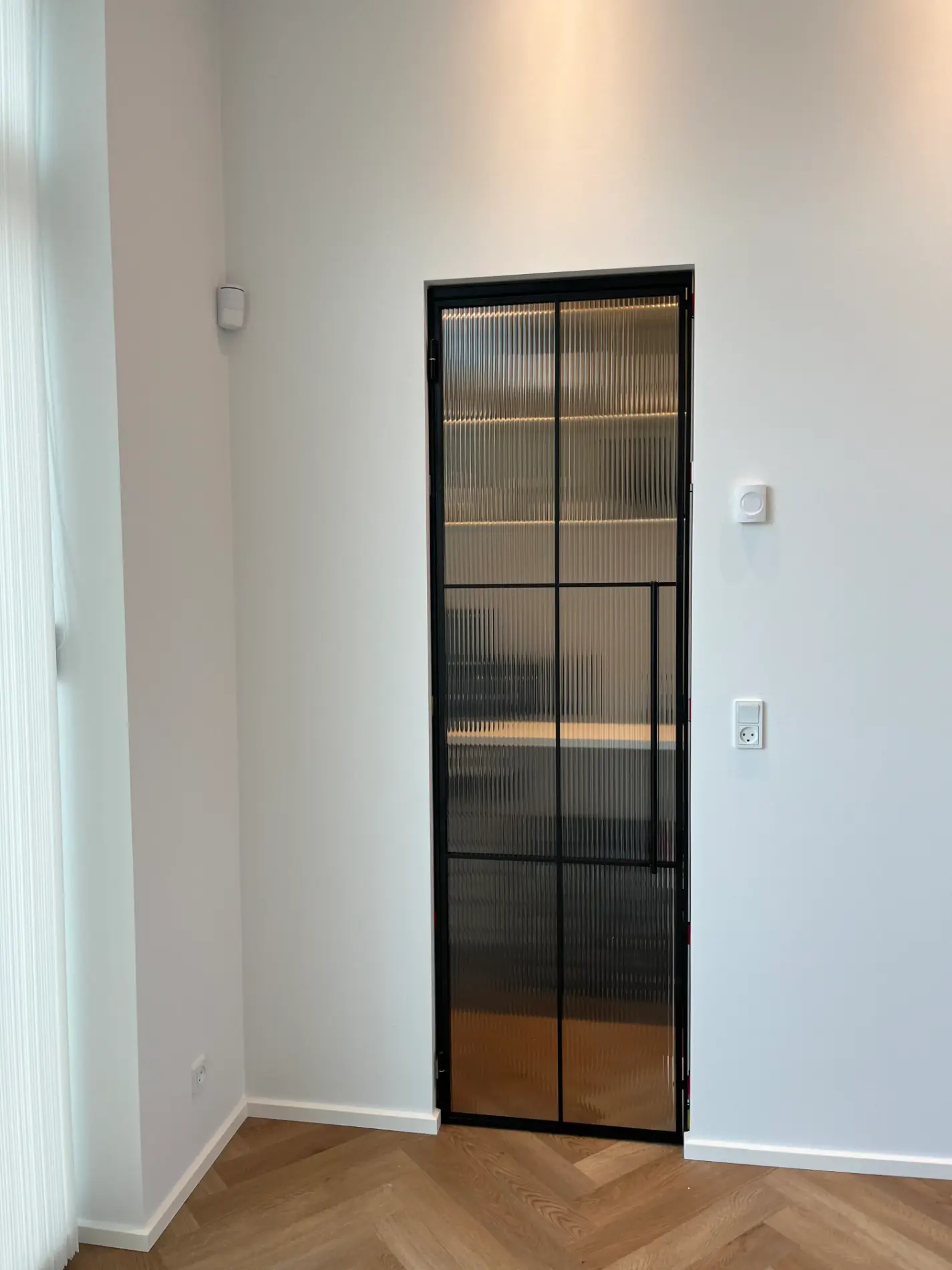 Sort metalrammedør med ribbet glas i en hvid væg og trægulv med sildebensmønster.
