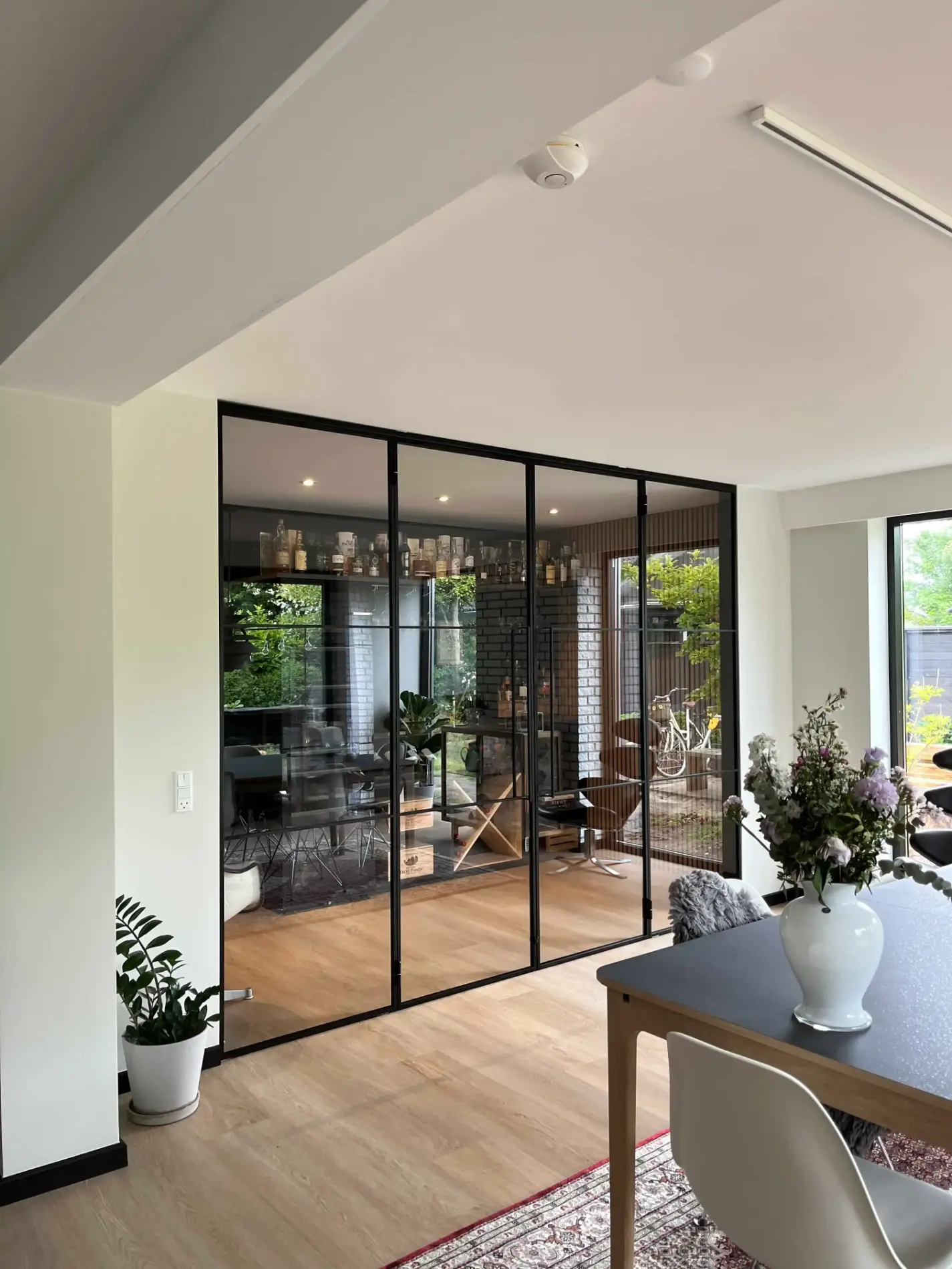 Moderne stue med glasvæg med sort ramme, trægulv, spisebord med hvid vase og blomster, samt udsigt til have og cykel.
