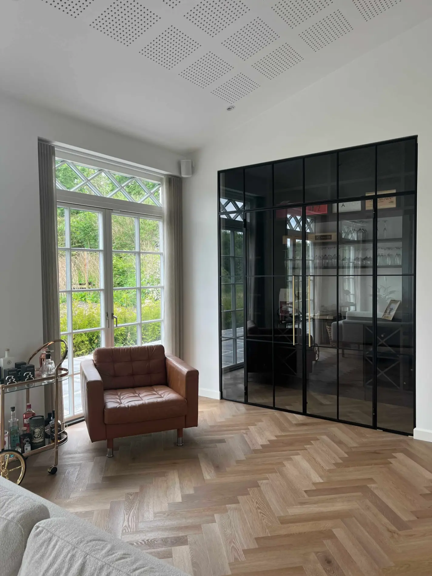 Moderne stue med lyst parketgulv, brun lænestol, glasvæg med sort ramme og store vinduer med udsigt til have.