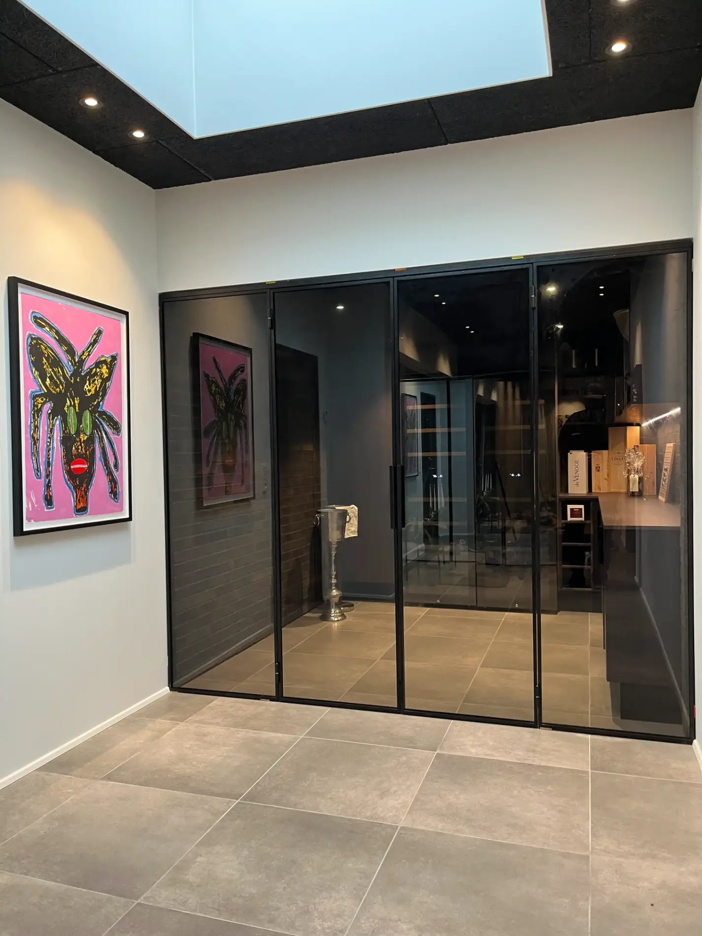 Moderne gang med store beige fliser, glasvægge og et farverigt kunstværk på en hvid væg under et loftvindue.