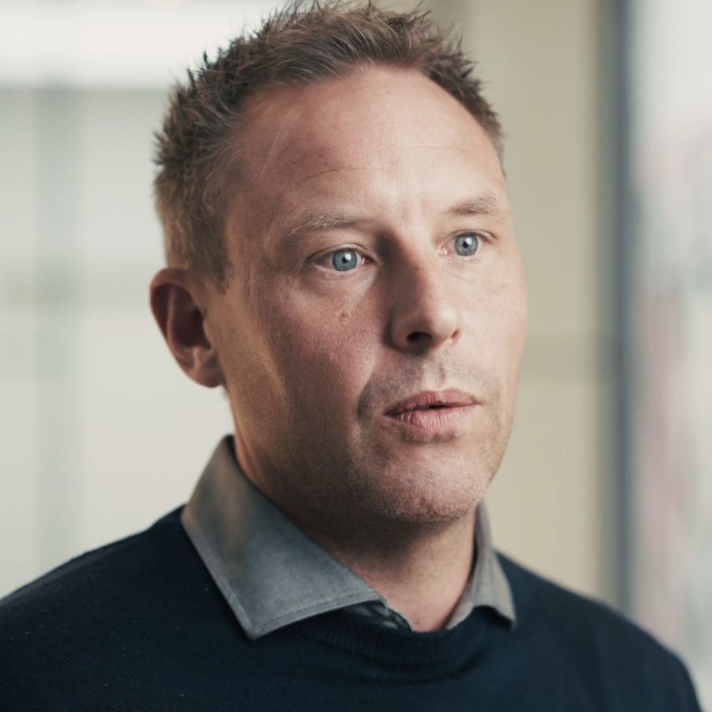 Portræt af en mand med kort, lysebrunt hår iført en grå skjorte og sort sweater, set mod en sløret baggrund.