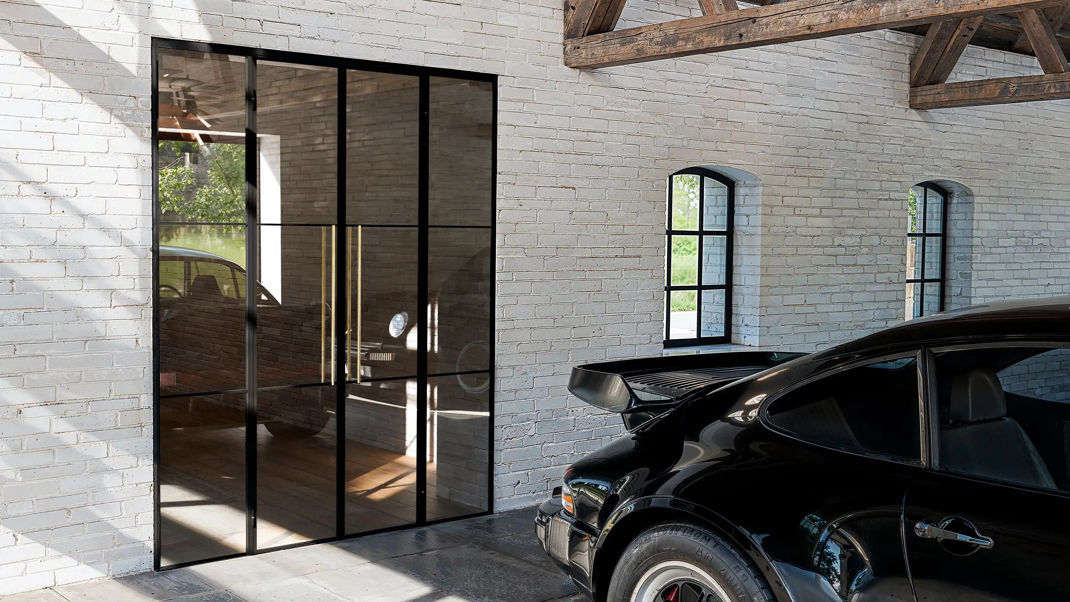 Sort sportsvogn parkeret i garage med hvidmalede murstensvægge og glasdør med sort ramme, der reflekterer bilens front.