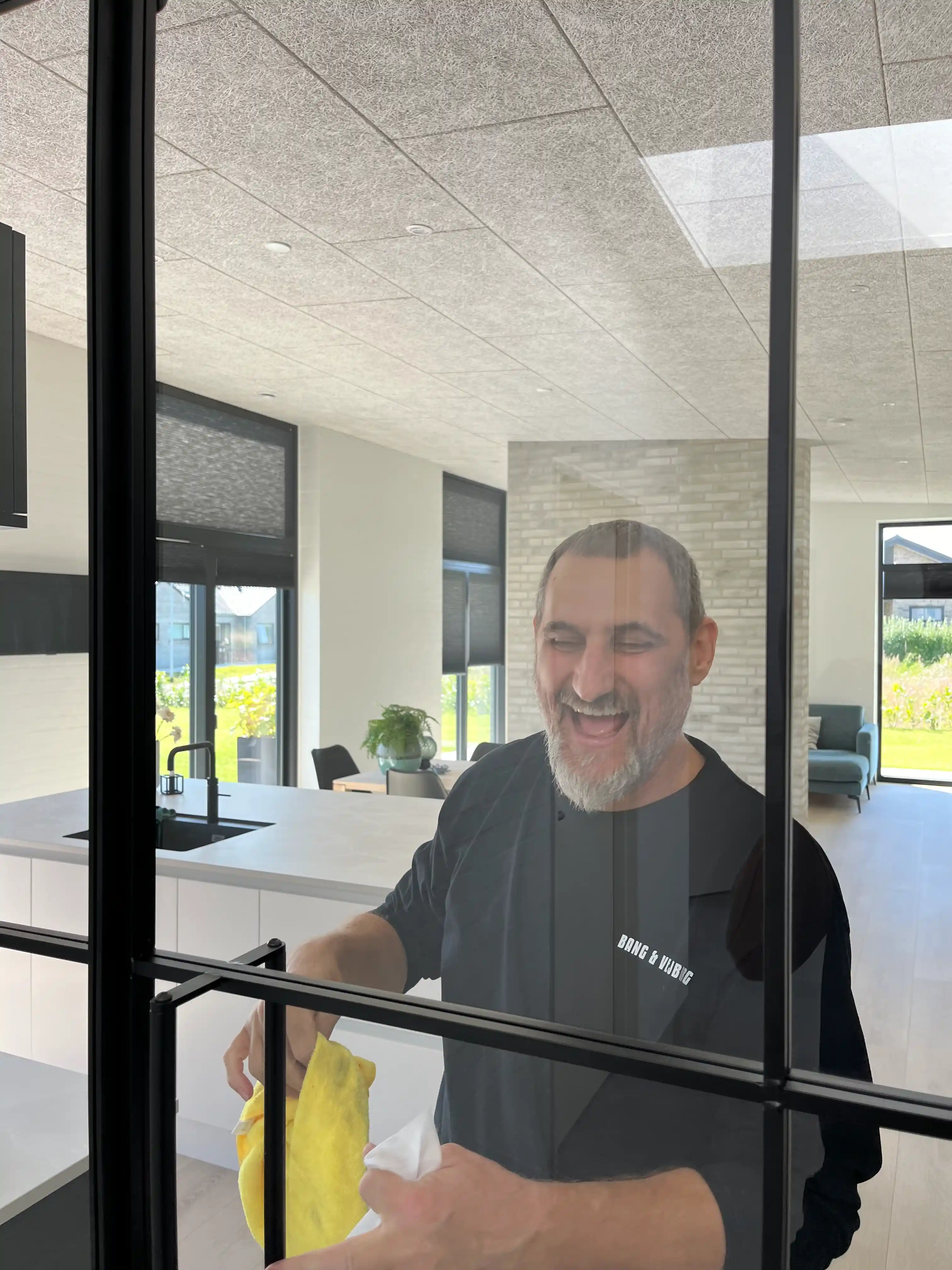 Smilende mand med grå skæg rengør et glasvindue med en gul klud i et lyst, moderne køkken.