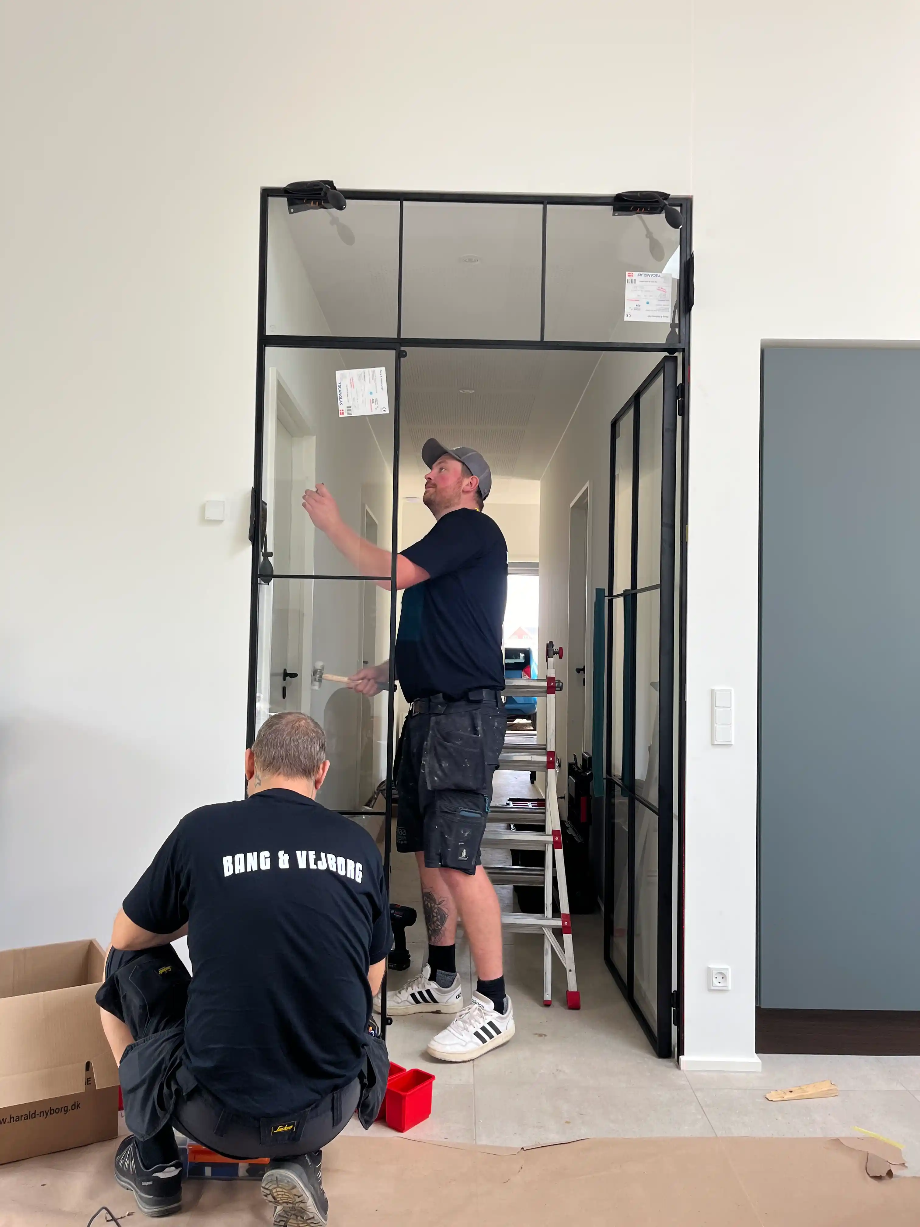 To håndværkere monterer en sort metal- og glasdør i en hvidt malet væg med et gråt dørblad ved siden af.