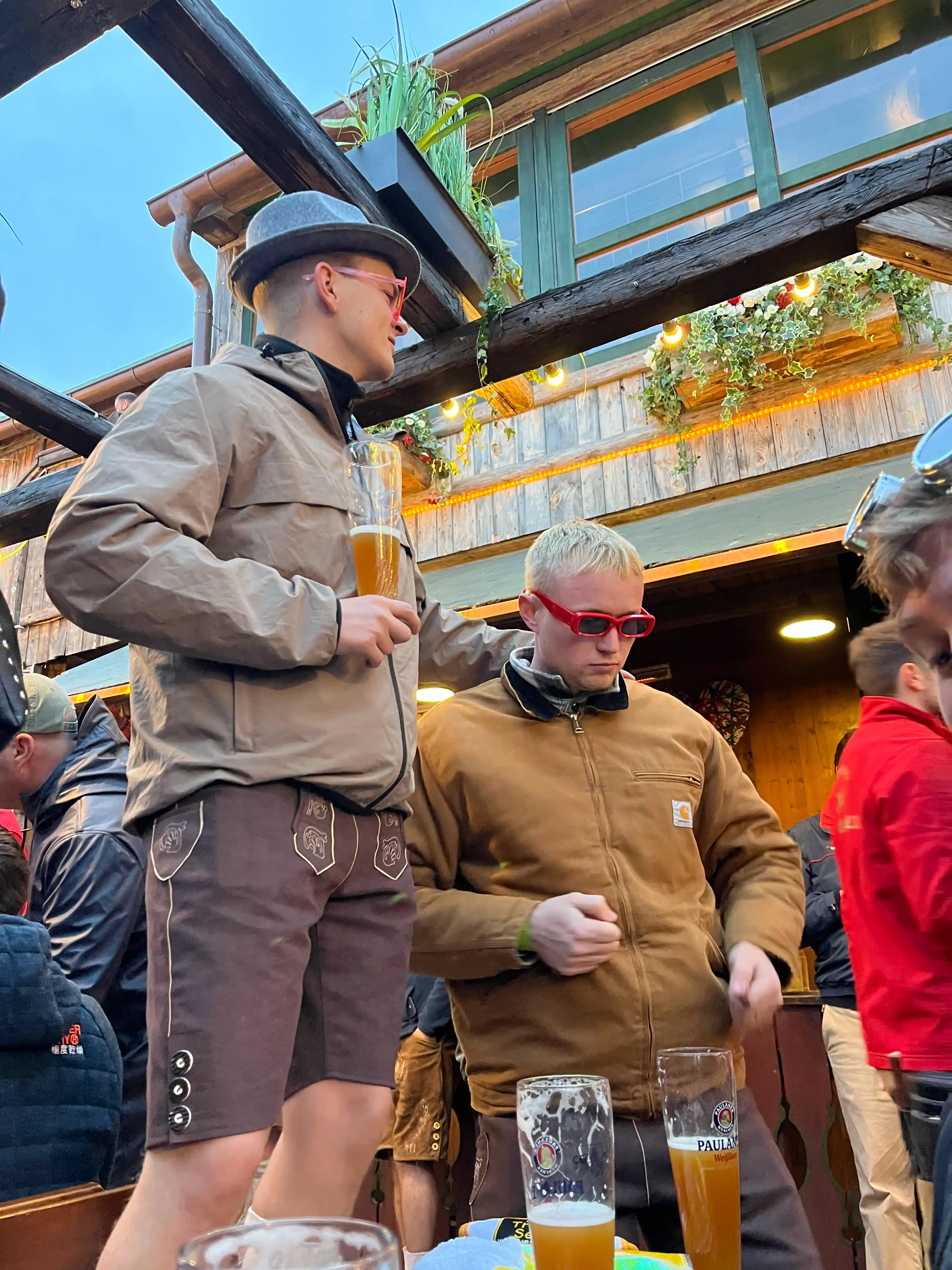 To mænd iført jakker og solbriller drikker øl ved et udendørs træsted med dekorative lyskæder.