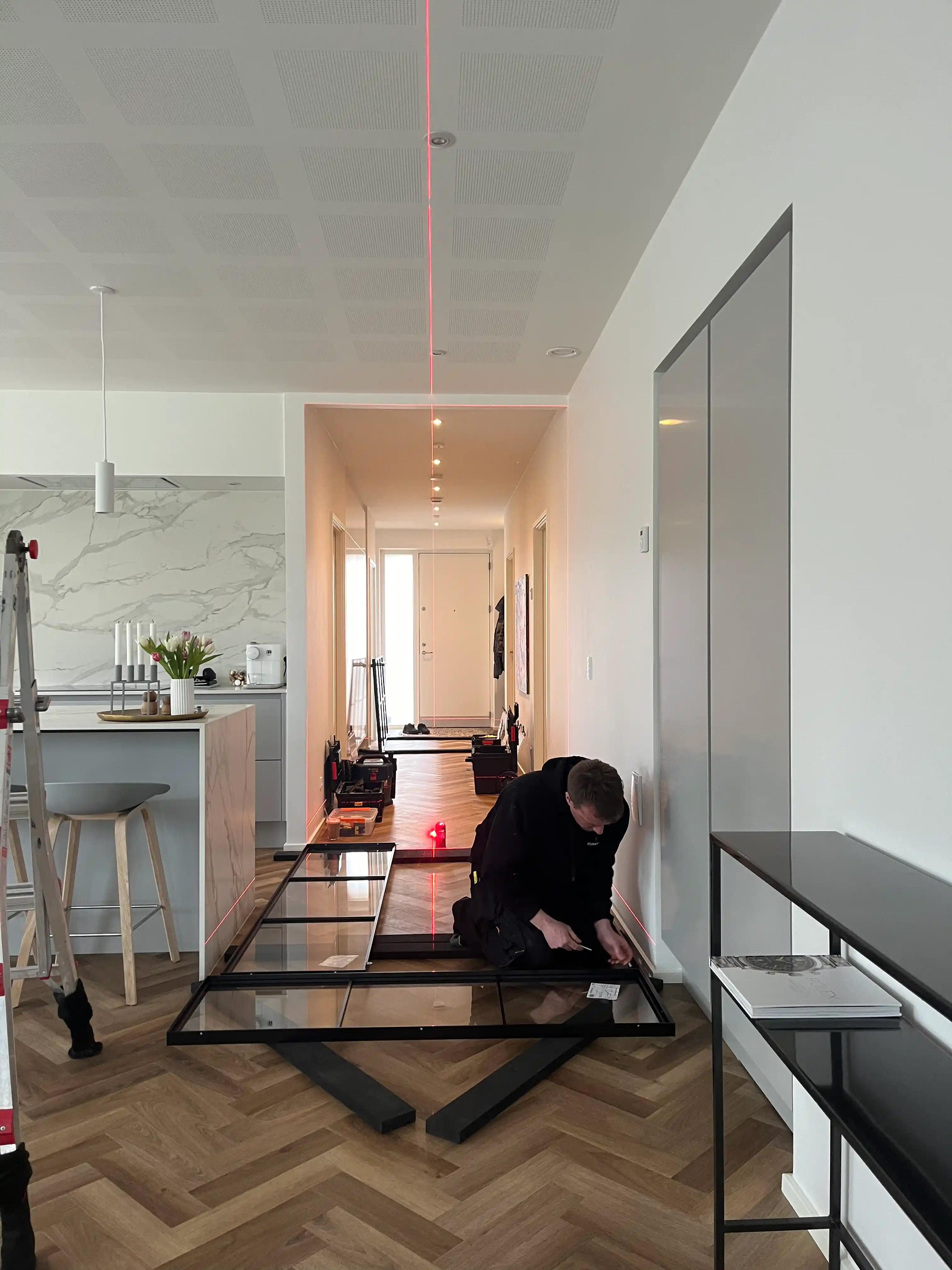 En håndværker monterer en glasdør på et stuegulv med parket, i en moderne lejlighed med laserlinien til opsætning.