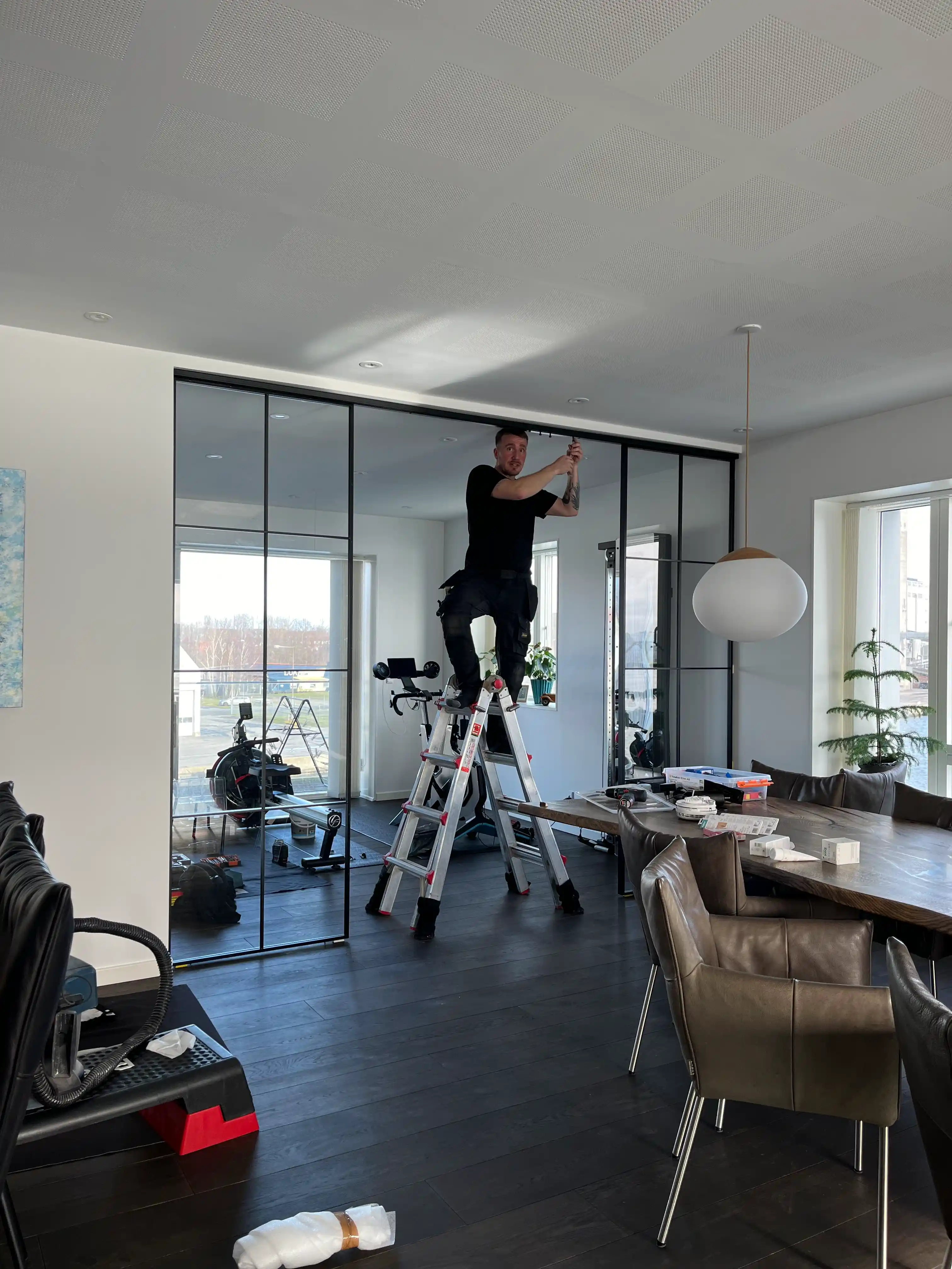 Mand står på en stige og arbejder på loftet i et moderne rum med mørkt gulv, glasvægge og brune læderstole omkring et bord.