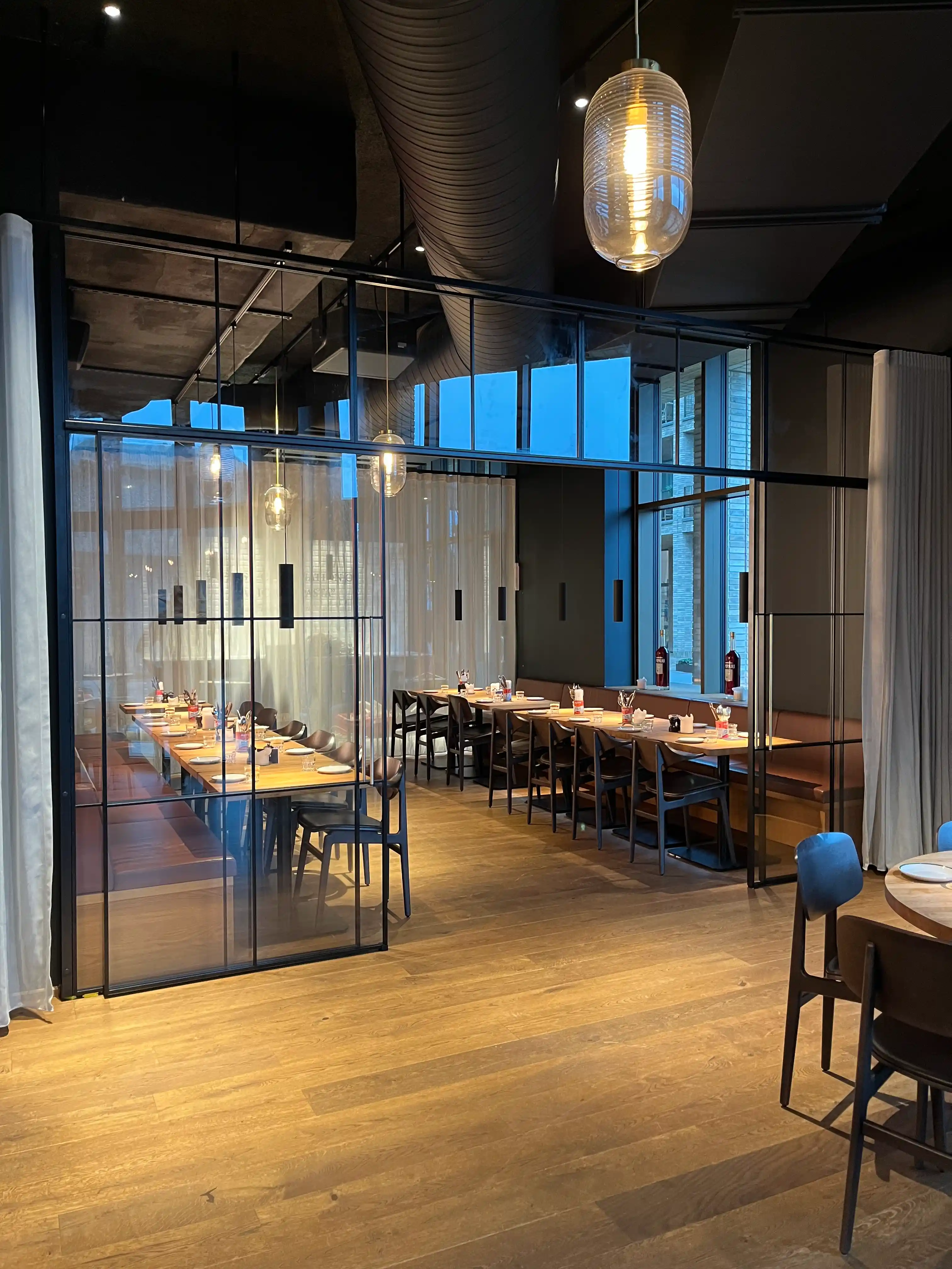 Moderne restaurant med træbord, sorte stole, glasvæg og hængende lamper.