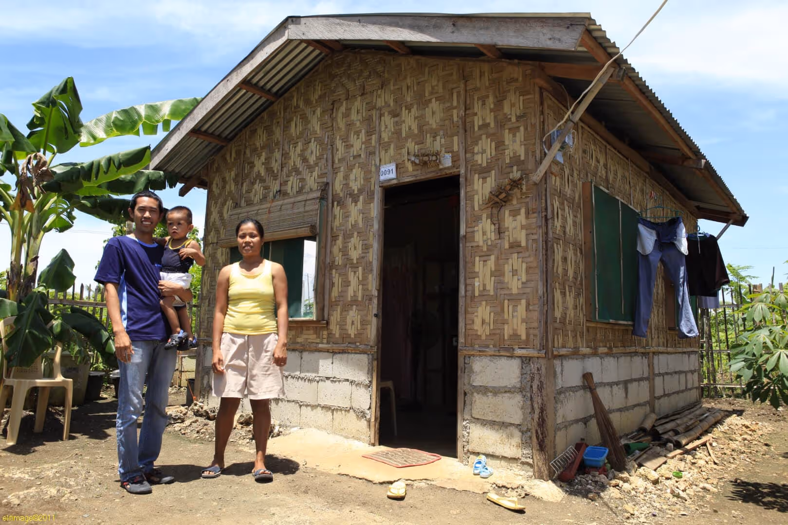 A family of two adults and a child standing in front of a small house with bamboo walls and a concrete base, with clothes hanging outside and banana plants nearby.