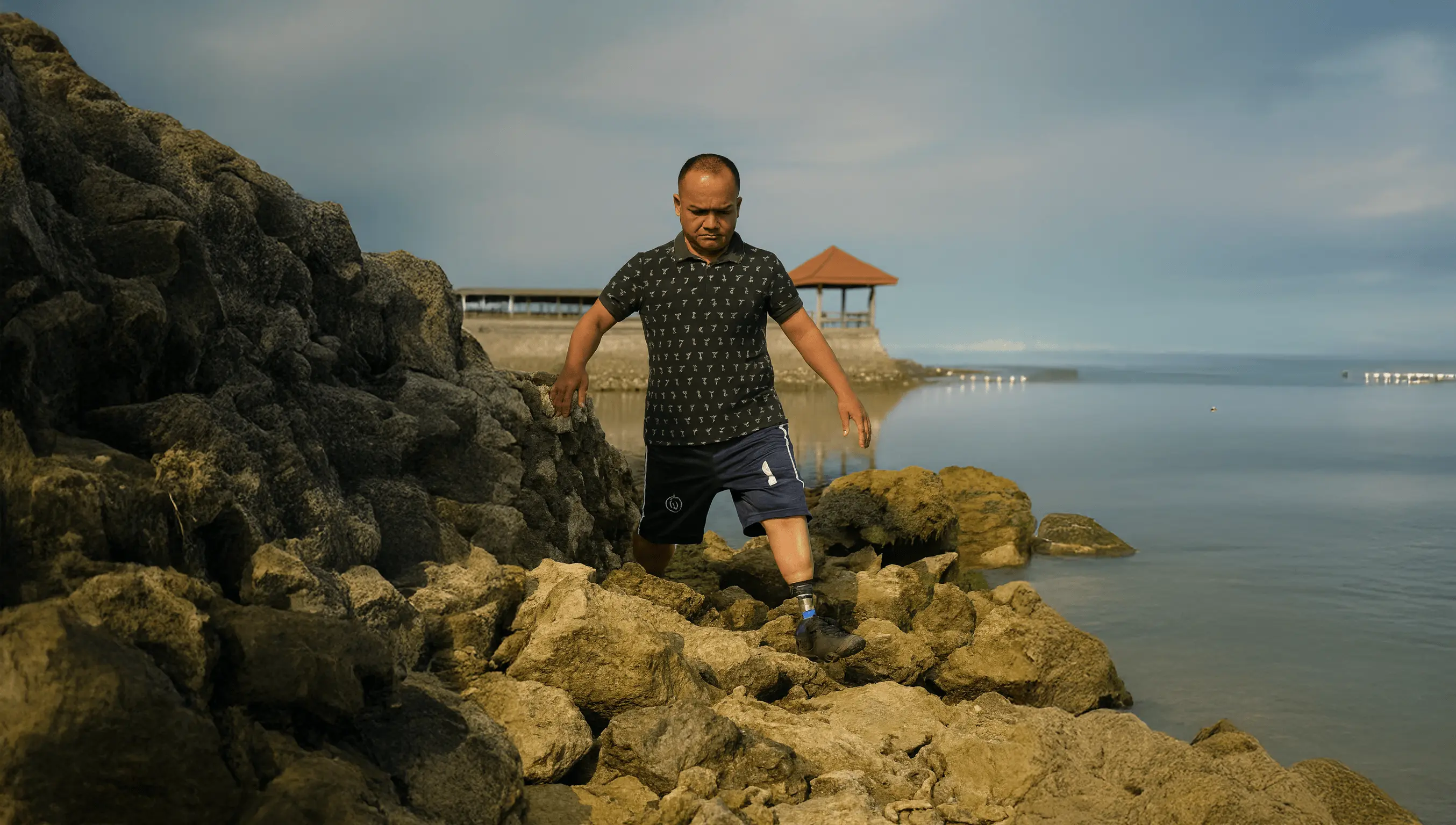 Man with a prosthetic leg walking carefully on rocky shore near calm water and a small pavilion in the background.