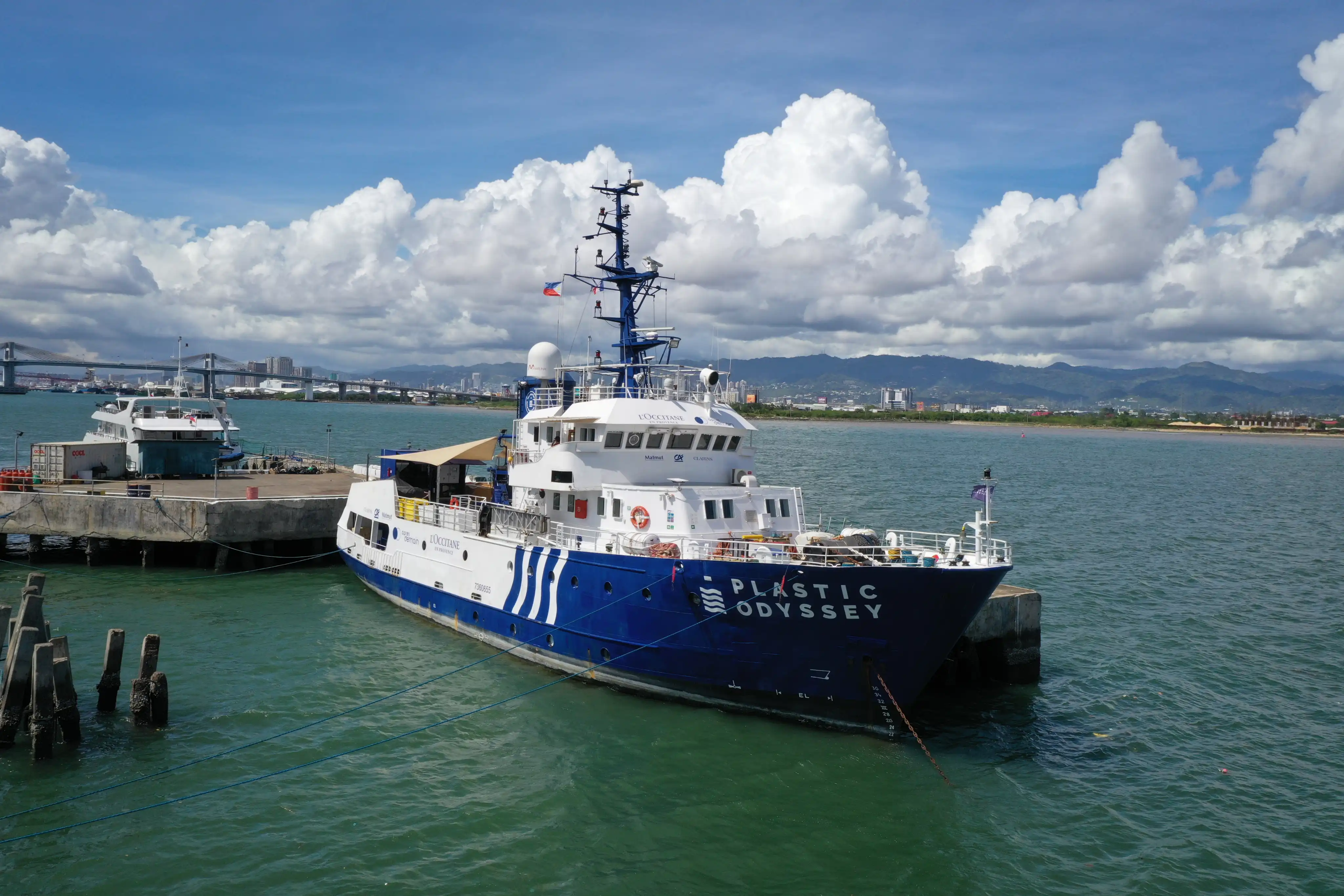 Research vessel Plastic Odyssey docked at a pier with mountains and cityscape in the background under a partly cloudy sky.