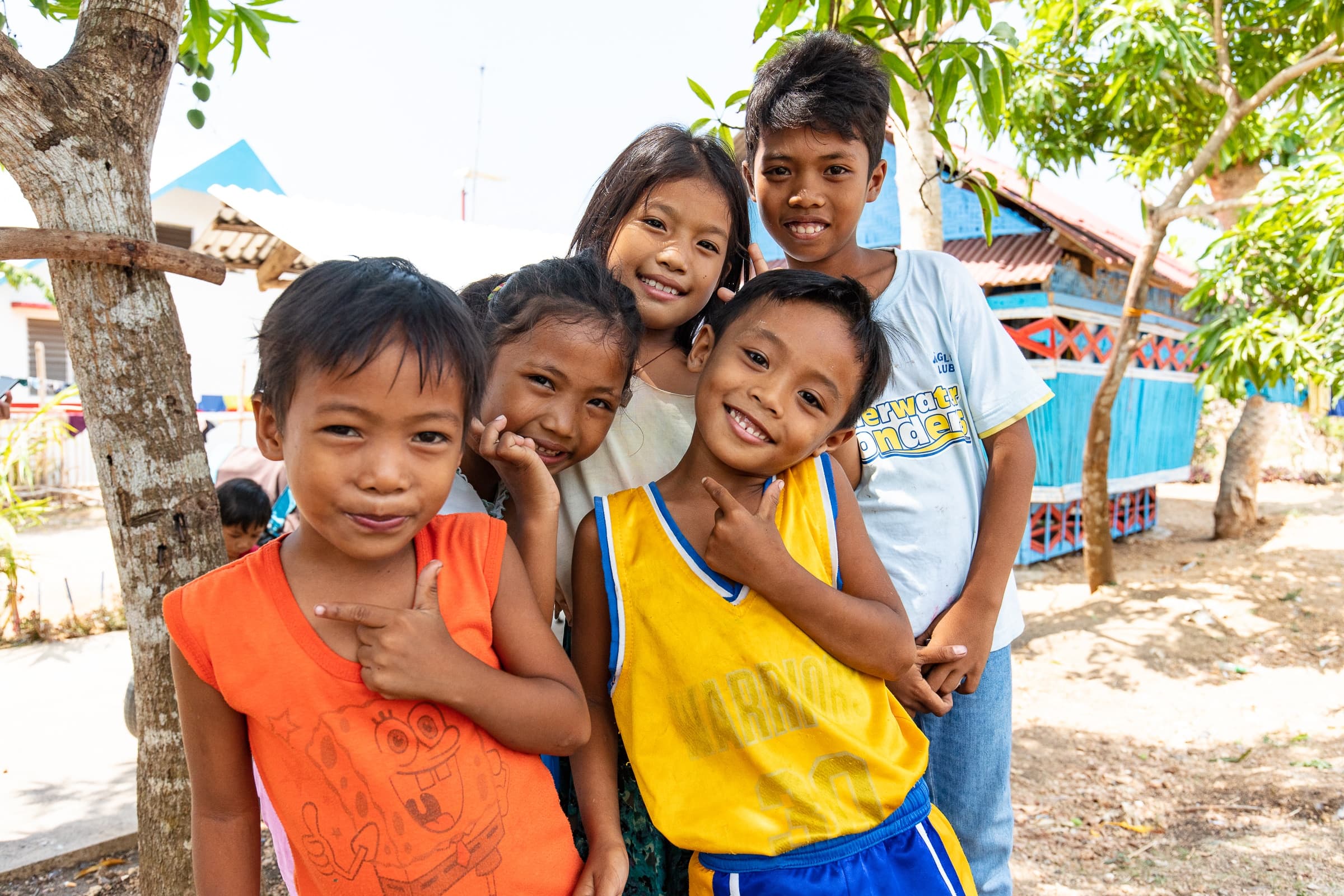 Group of five smiling children posing outdoors with trees and colorful buildings in the background.