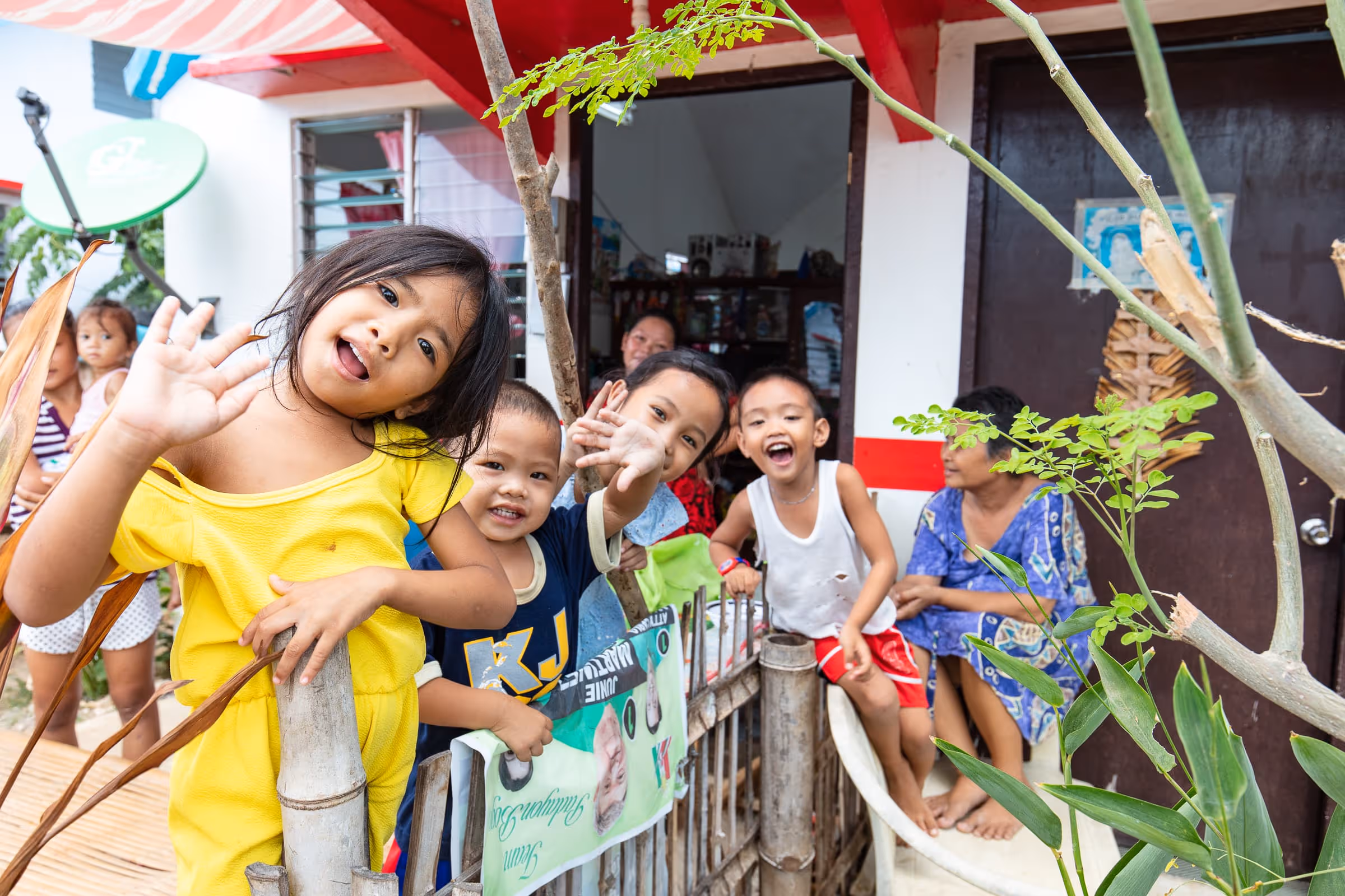 Group of smiling children greeting and waving near a bamboo fence outside a house with adults sitting in the background.