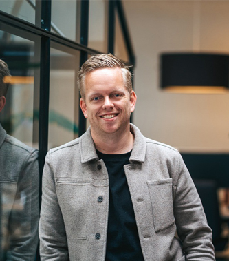 Smiling man with light brown hair wearing a gray jacket and black shirt, standing indoors next to a glass window.