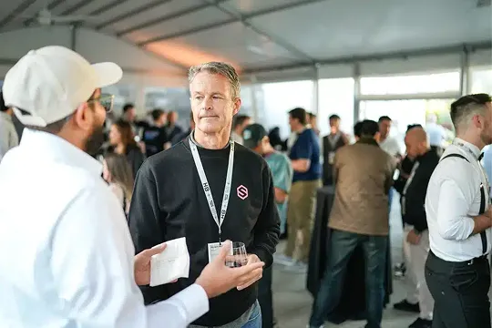 Two men conversing at a social event indoors with blurred groups of people mingling in the background.