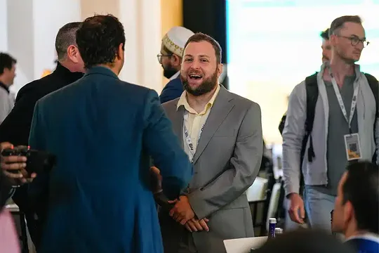 Group of men engaged in conversation at a professional indoor event, one man in a grey suit is smiling.