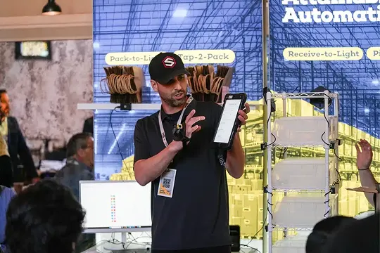 Man wearing a black cap and shirt demonstrating a handheld device in front of a large screen with logistics and automation terms.